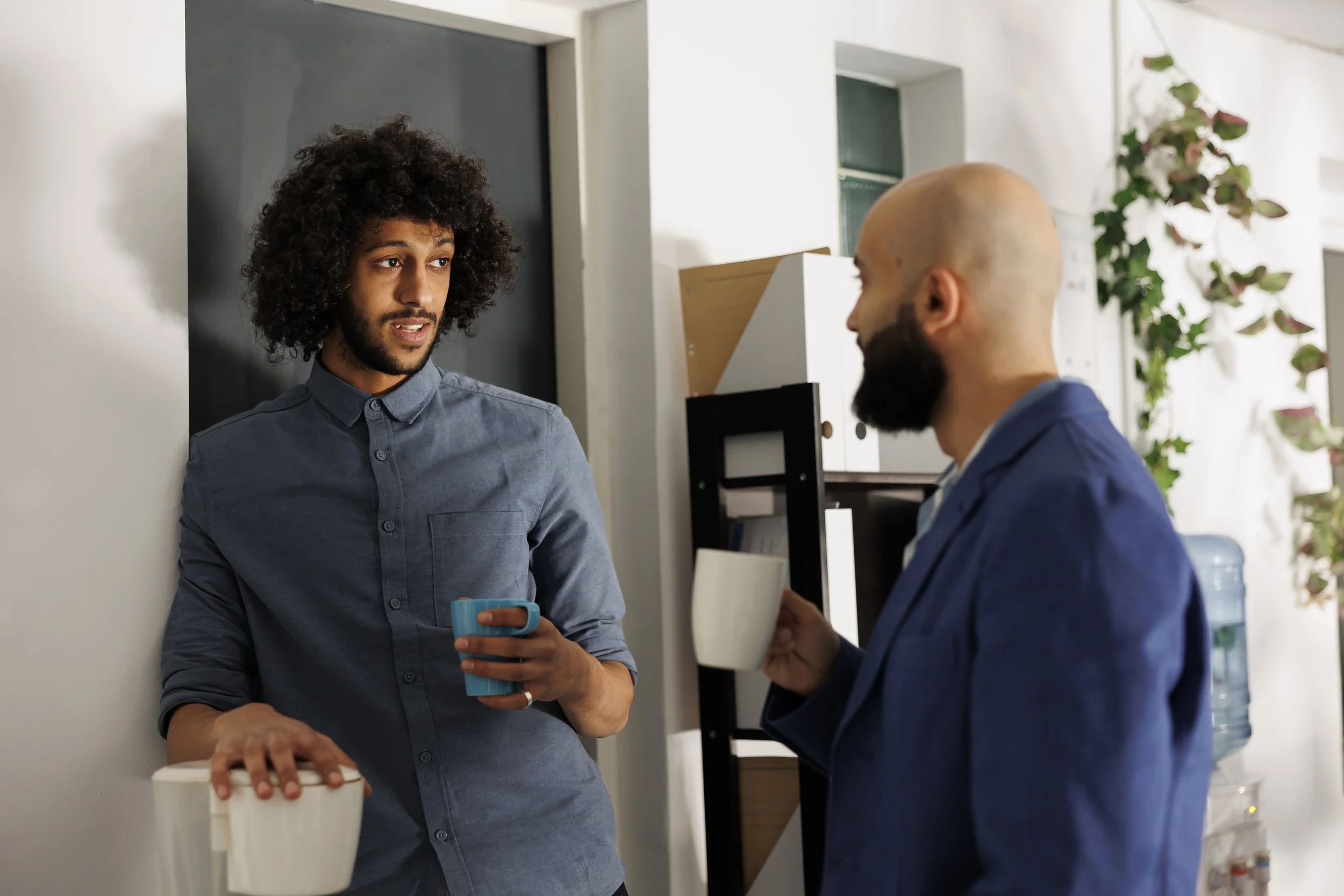 Two men are having a conversation in an office. One man is holding a blue mug and appears to be listening while the other man is speaking. The office background has a blackboard, some shelves with folders, and a plant.