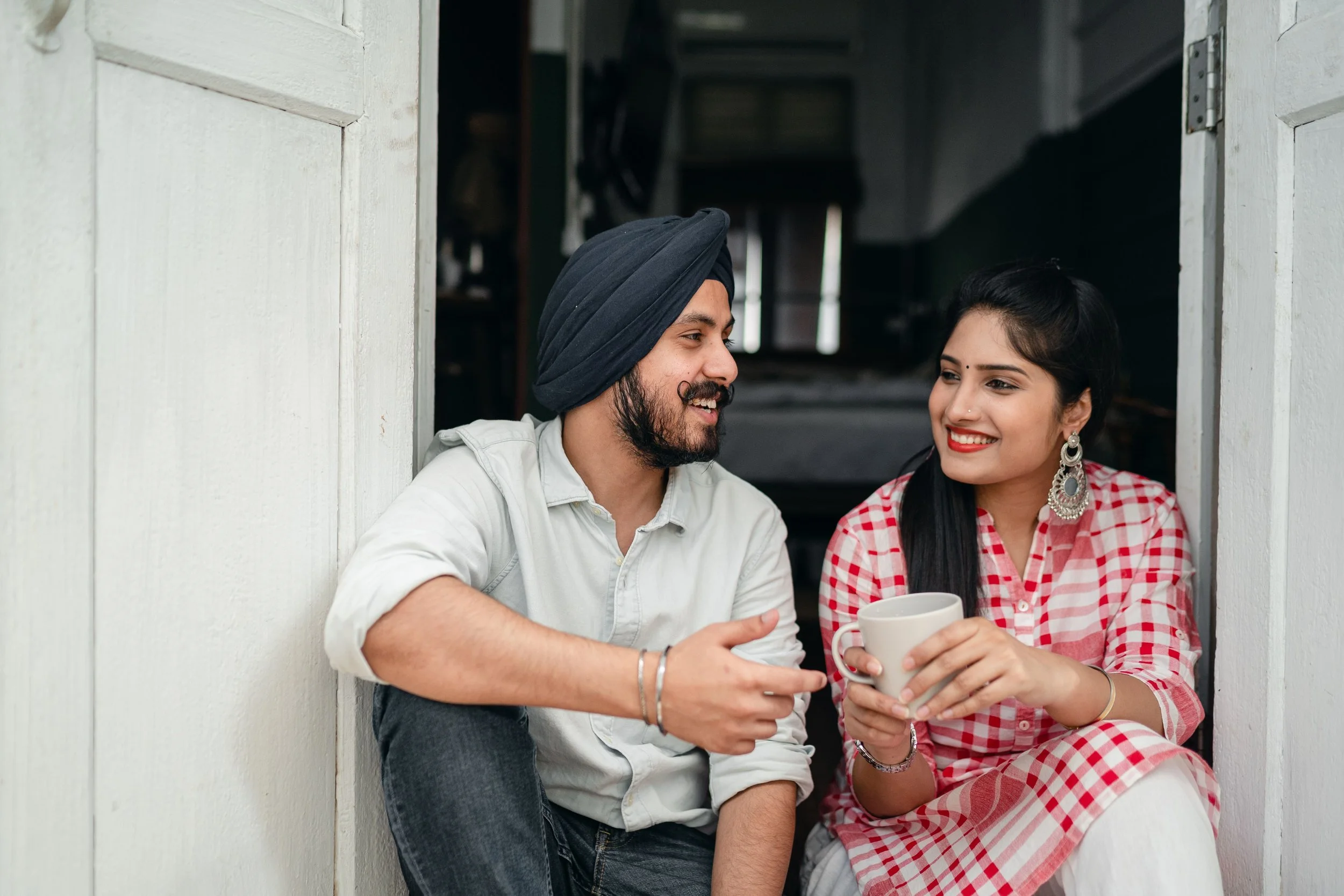 A man with a beard and a woman with long dark hair sitting at a white wooden window frame, smiling and holding coffee mugs, in a cozy indoor setting.