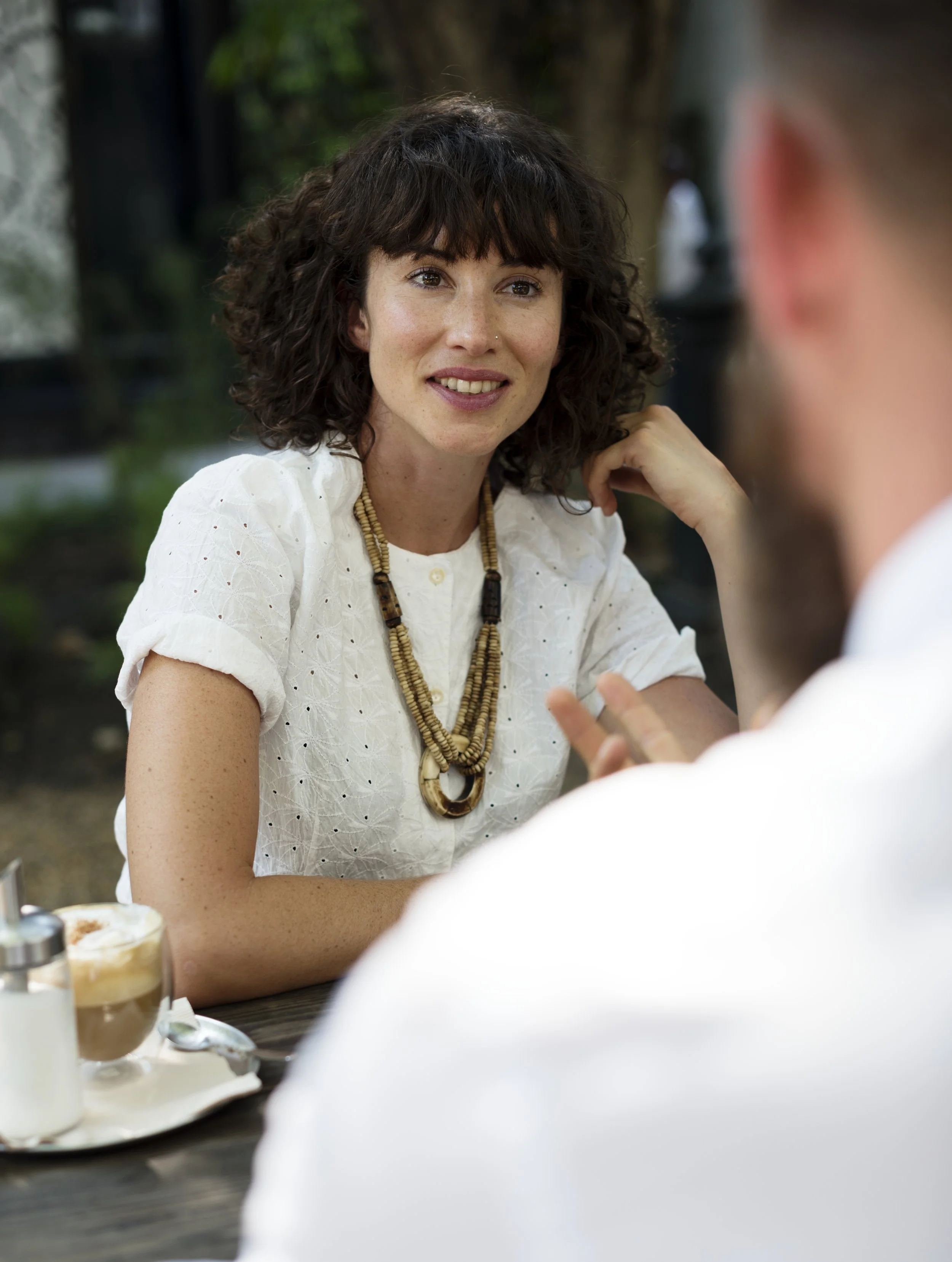 A woman with dark curly hair, wearing a white eyelet blouse and beaded necklace, is sitting at an outdoor table and smiling while talking to someone.