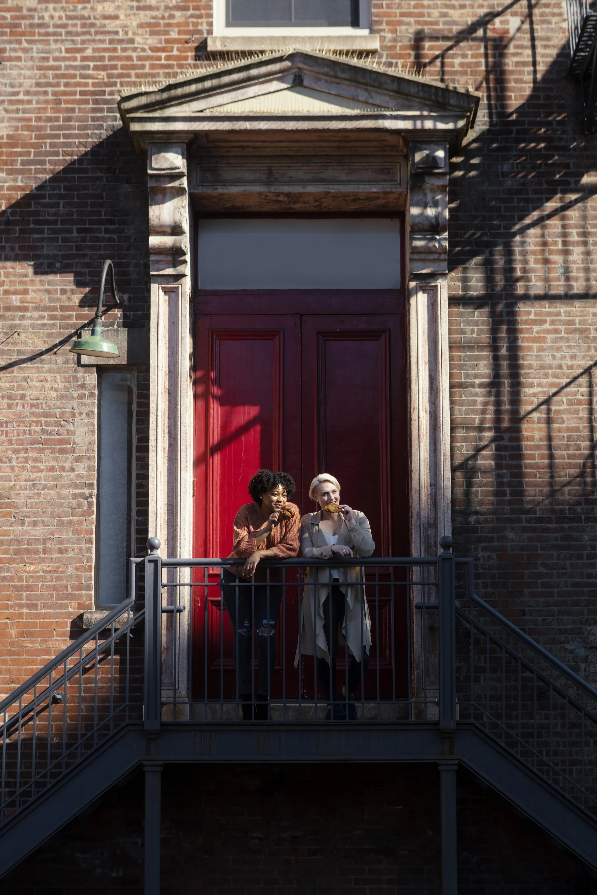 Two women standing on a small balcony in front of a large red door with brick wall background, eating pizza. One woman has curly black hair and a brown sweater, the other has short blonde hair and a beige trench coat.