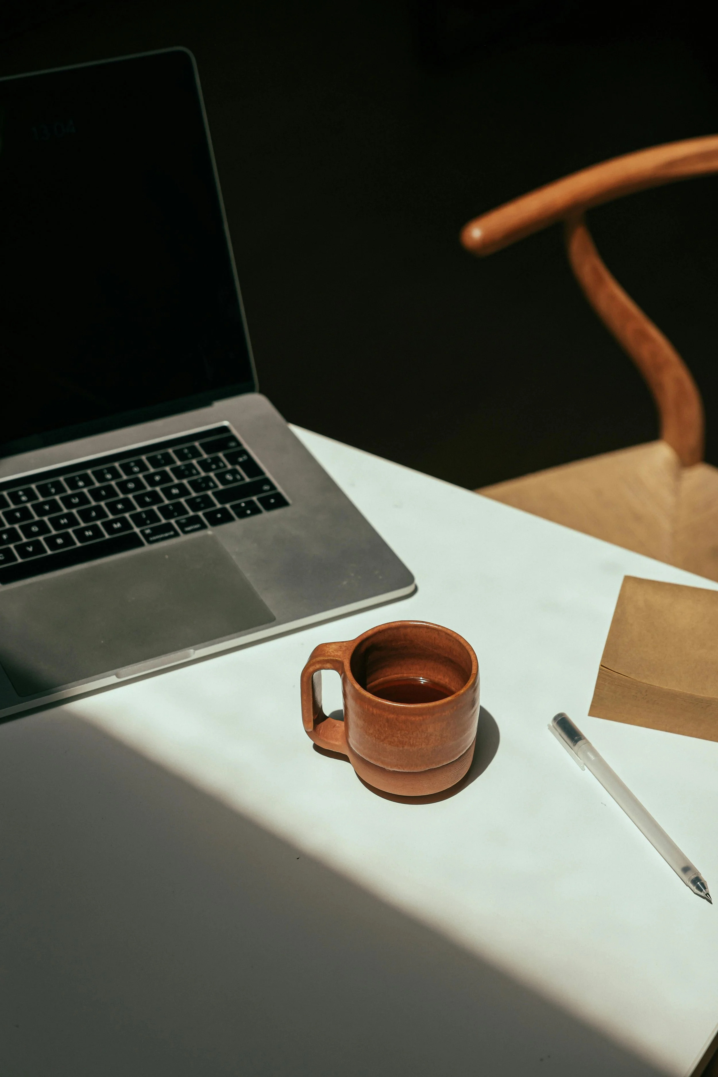 A laptop on a white desk, a ceramic mug filled with a dark beverage, a transparent pen, and brown paper on the desk, with a black background and a wooden chair partially visible.