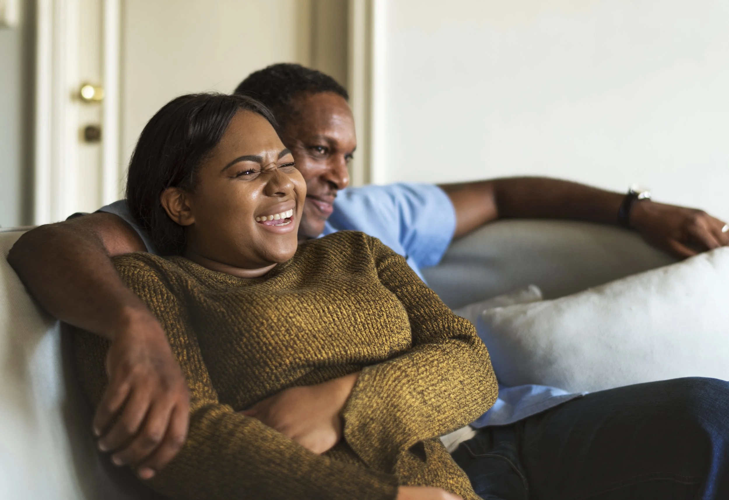 A woman with long dark hair and a brown sweater is smiling with her eyes closed, sitting on a couch with a man in a light blue shirt, both looking happy and relaxed.