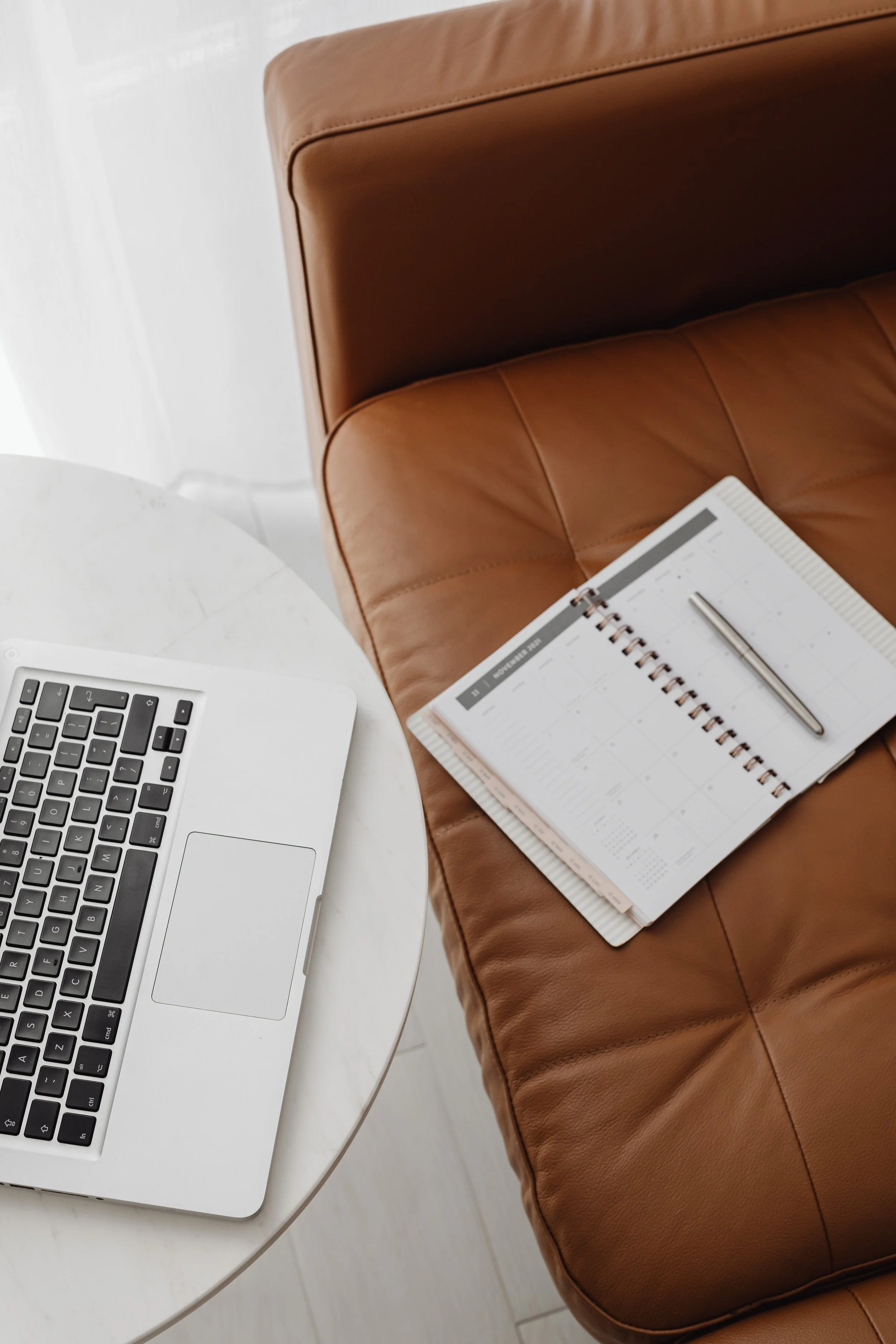 Computer keyboard, white table, brown leather couch with open planner and pen on it, part of a white curtain in the background.