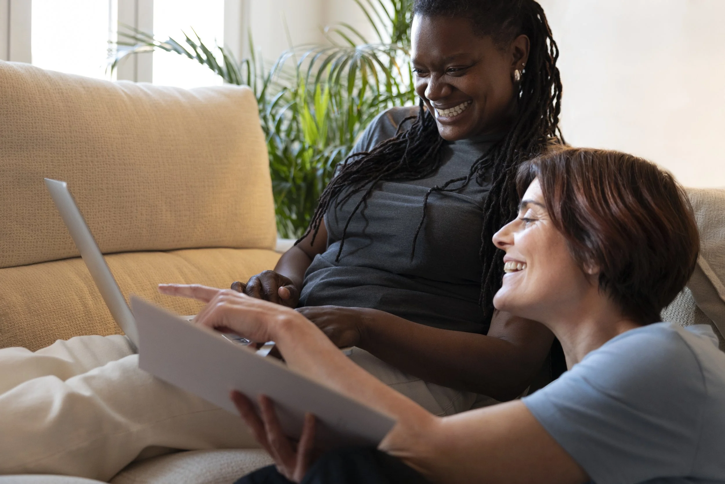 Two women sitting on a couch, smiling and looking at a laptop together in a bright living room with plants in the background.