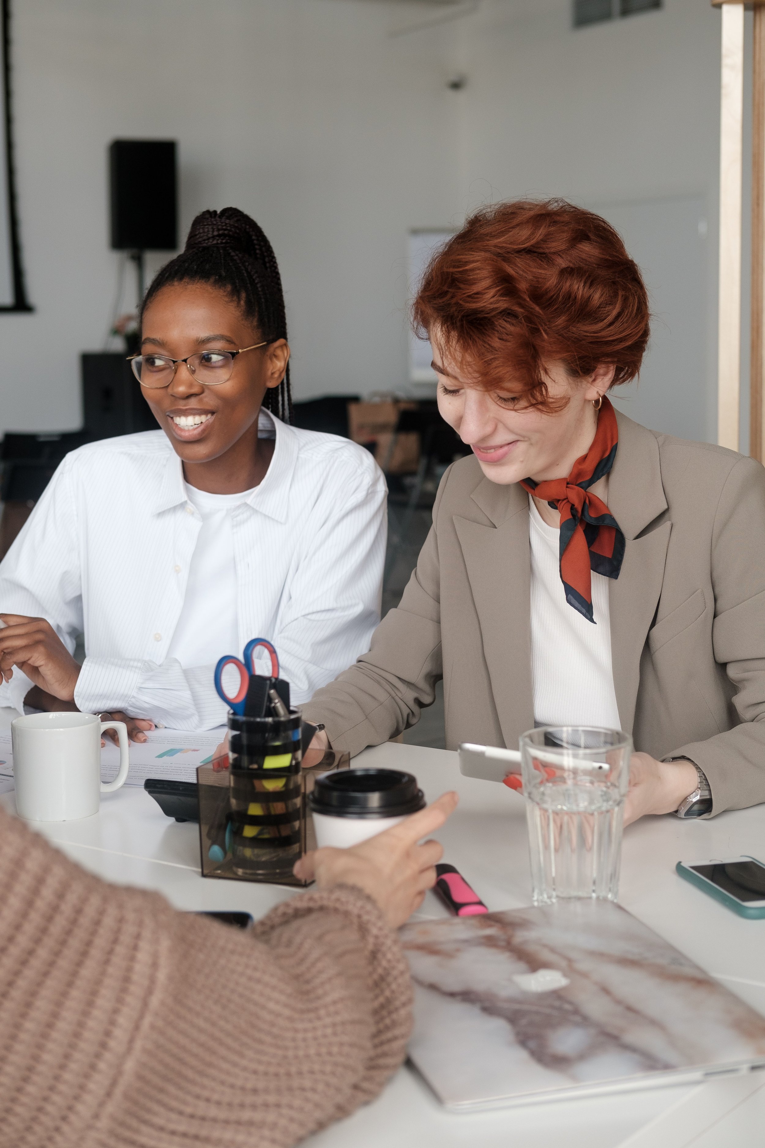 Two women sitting at a table engaged in conversation. One woman has dark skin, glasses, and braided hair tied up, and is smiling. The other woman has lighter skin, short wavy red hair, and is wearing a tan blazer with a red and blue scarf, and is looking down. On the table are various items including a white mug, a pair of scissors, a black container with pens, a glass of water, and a small cup with a lid. The background shows a plain white wall and some furniture.