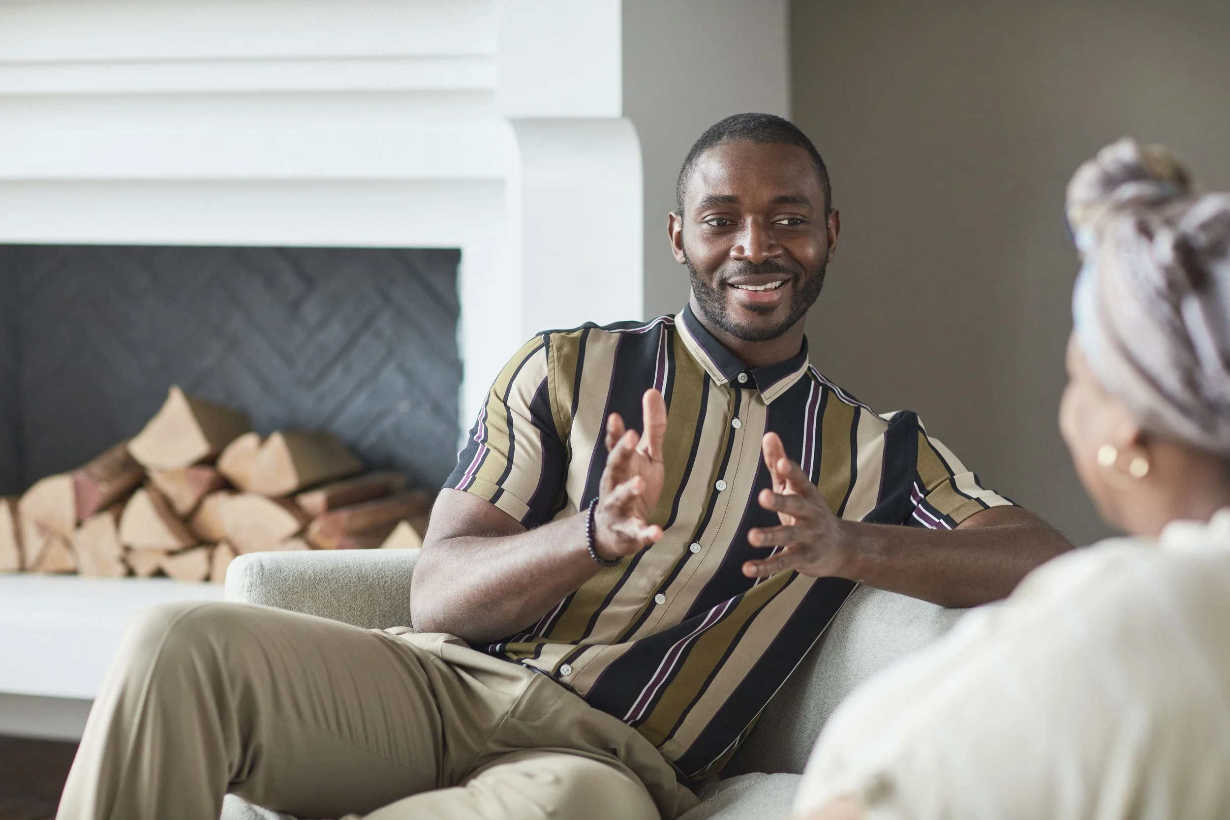 A man with short dark hair, a beard, and a striped shirt is speaking to a woman with glasses and a patterned headwrap in a cozy indoor setting.