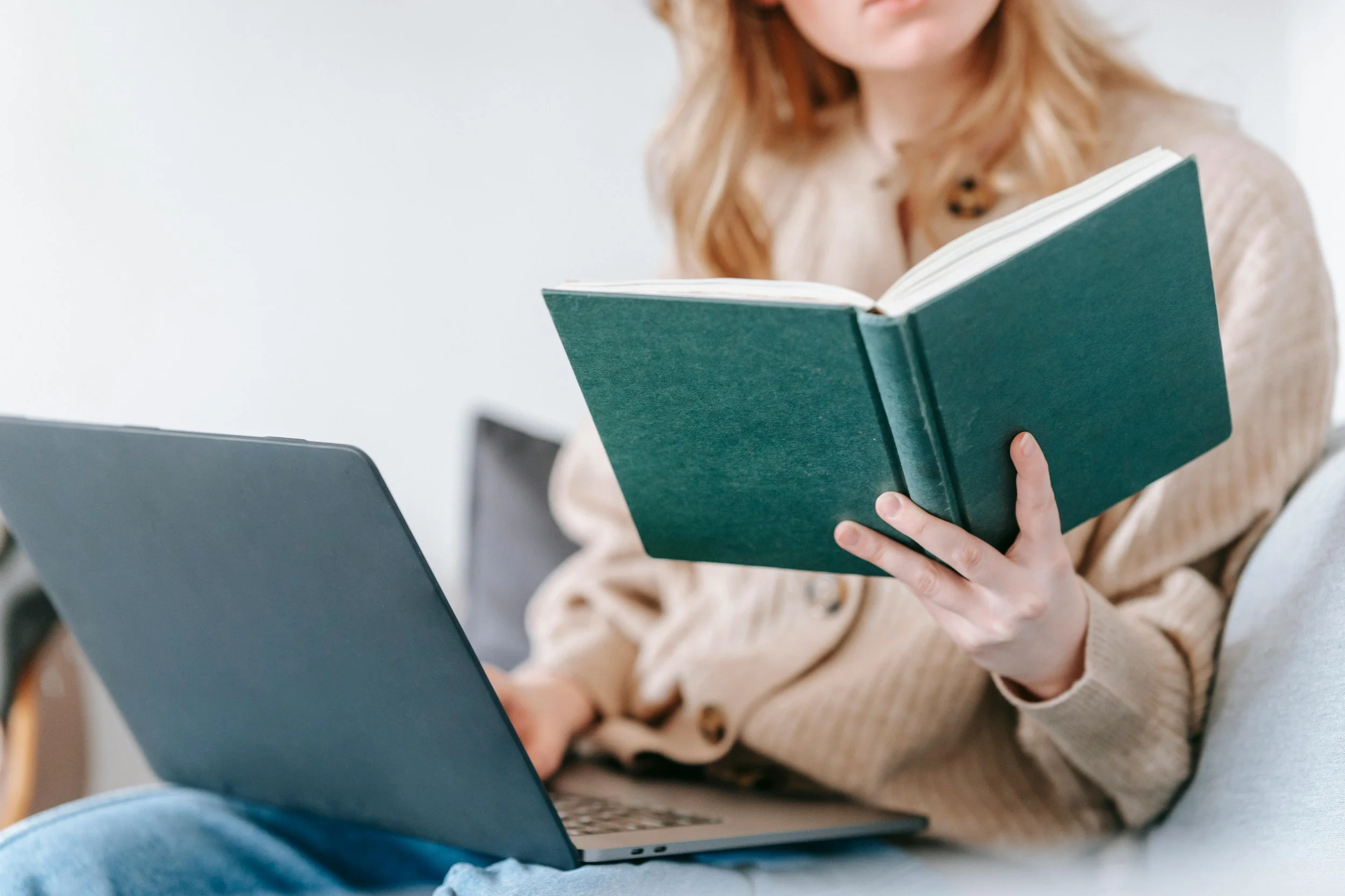 A woman with red hair reading a green hardcover book while sitting at a desk with a laptop.