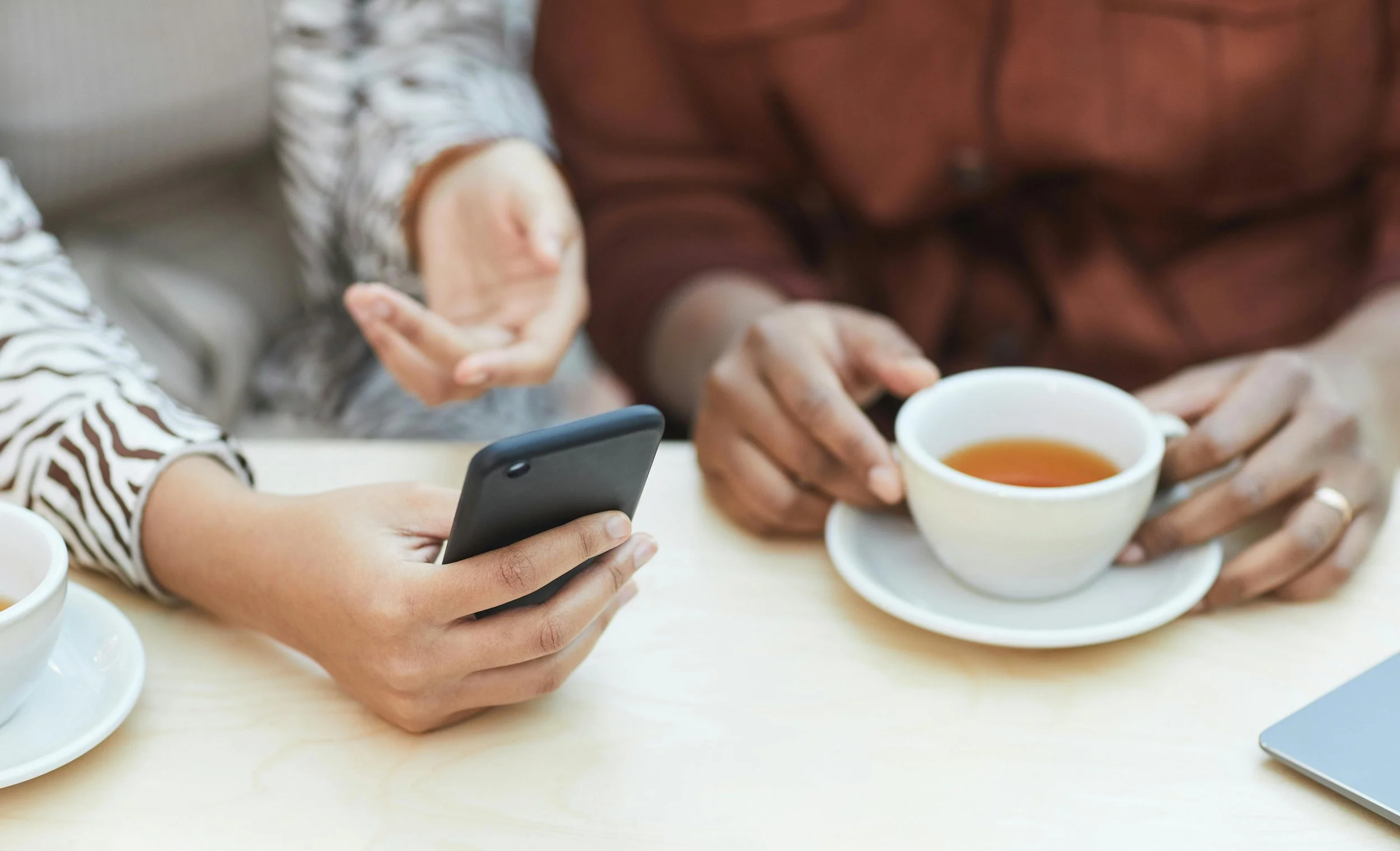 Person holding a smartphone in their left hand, and a person with glasses and a brown shirt holding a cup of tea with their right hand in the background. There are multiple white cups of tea on the table.