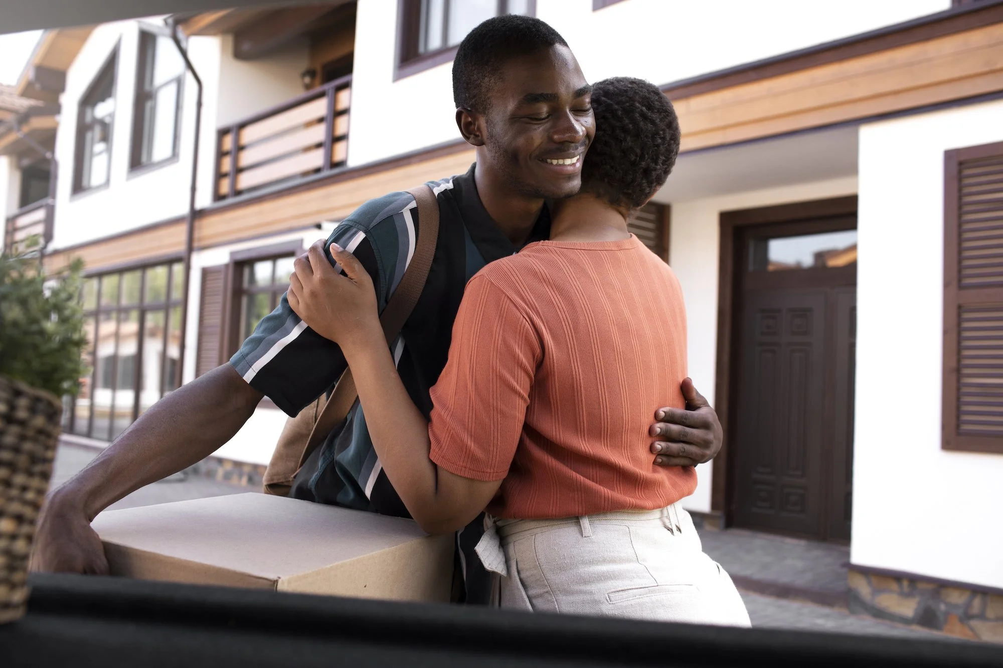 A young man and woman hugging in front of a house. The man is smiling with eyes closed, wearing a striped shirt and a backpack. The woman has short curly hair, is facing away, and is wearing a peach-colored top and beige pants.