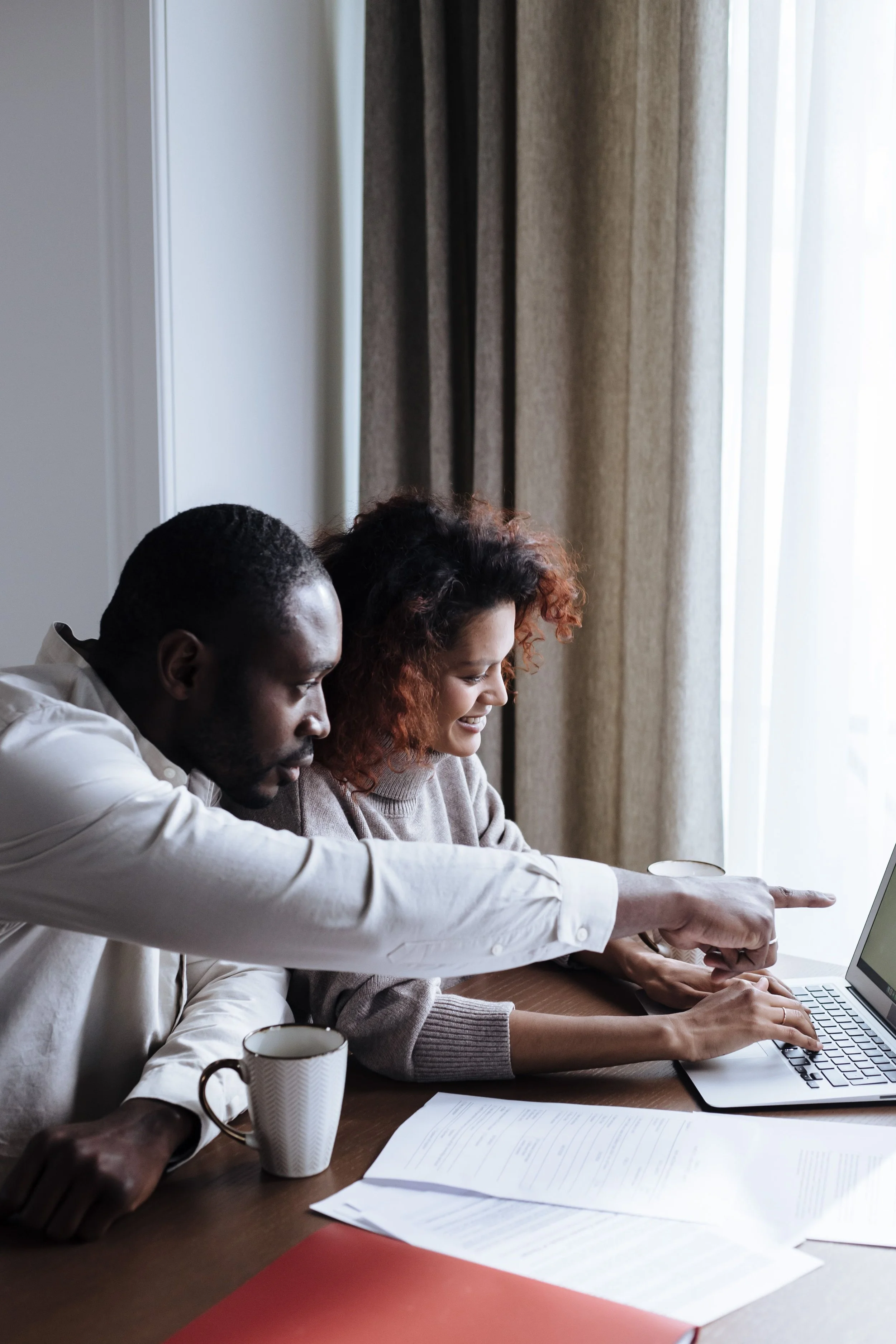 Two people, a man and a woman, are sitting at a table looking at a laptop screen. The man is pointing at the screen, and the woman is smiling. There are papers, a coffee mug, and a red folder on the table. They are near a window with curtains.