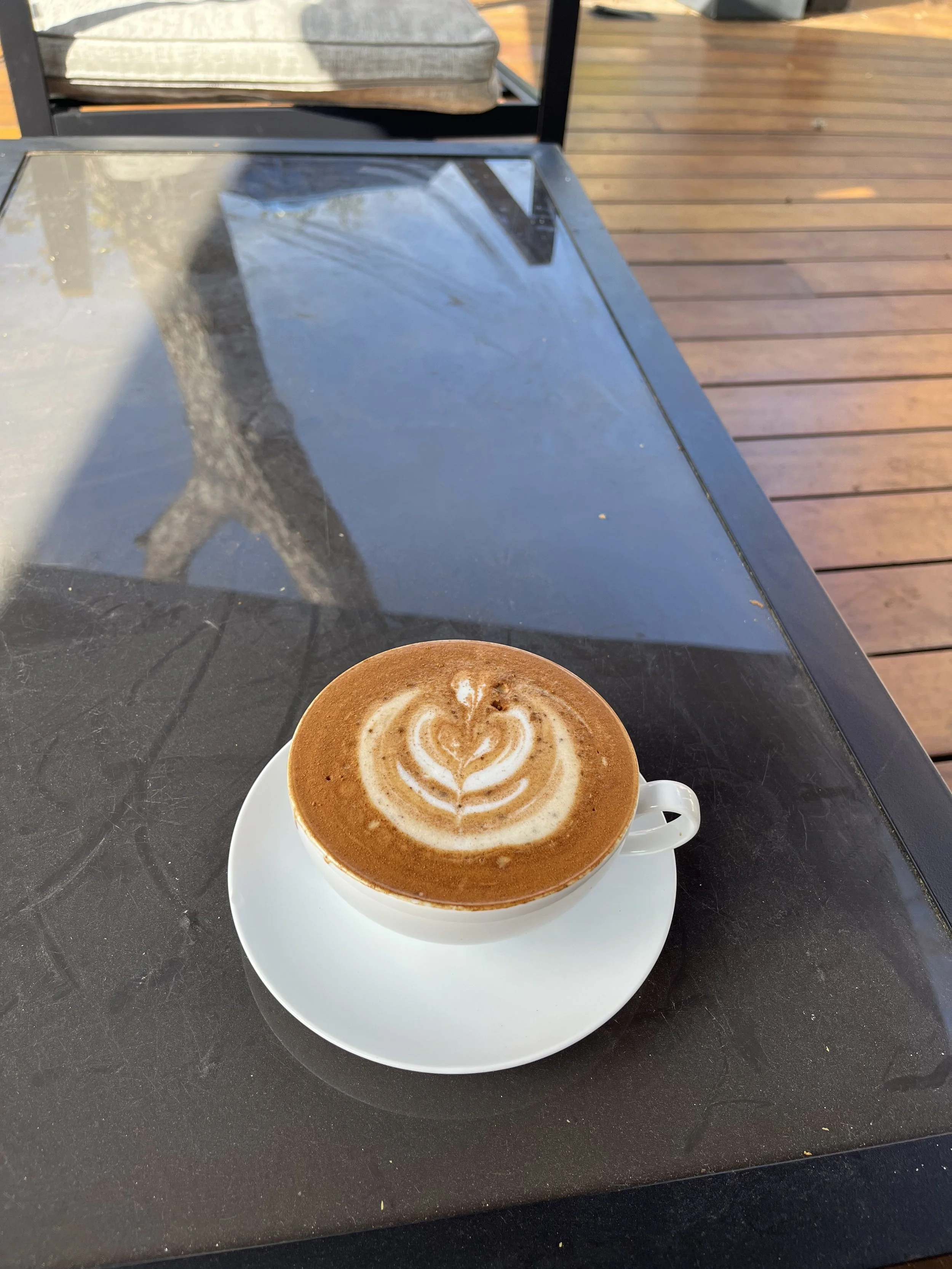 A white cup of cappuccino with latte art on top sits on a white saucer on a black table outside. In the background, there is a wooden deck and an outdoor chair with a cushion.
