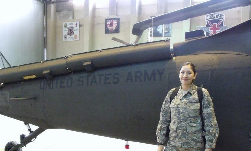 A woman in a camouflage military uniform standing inside an aircraft hangar next to a military helicopter with 'United States Army' painted on its side.
