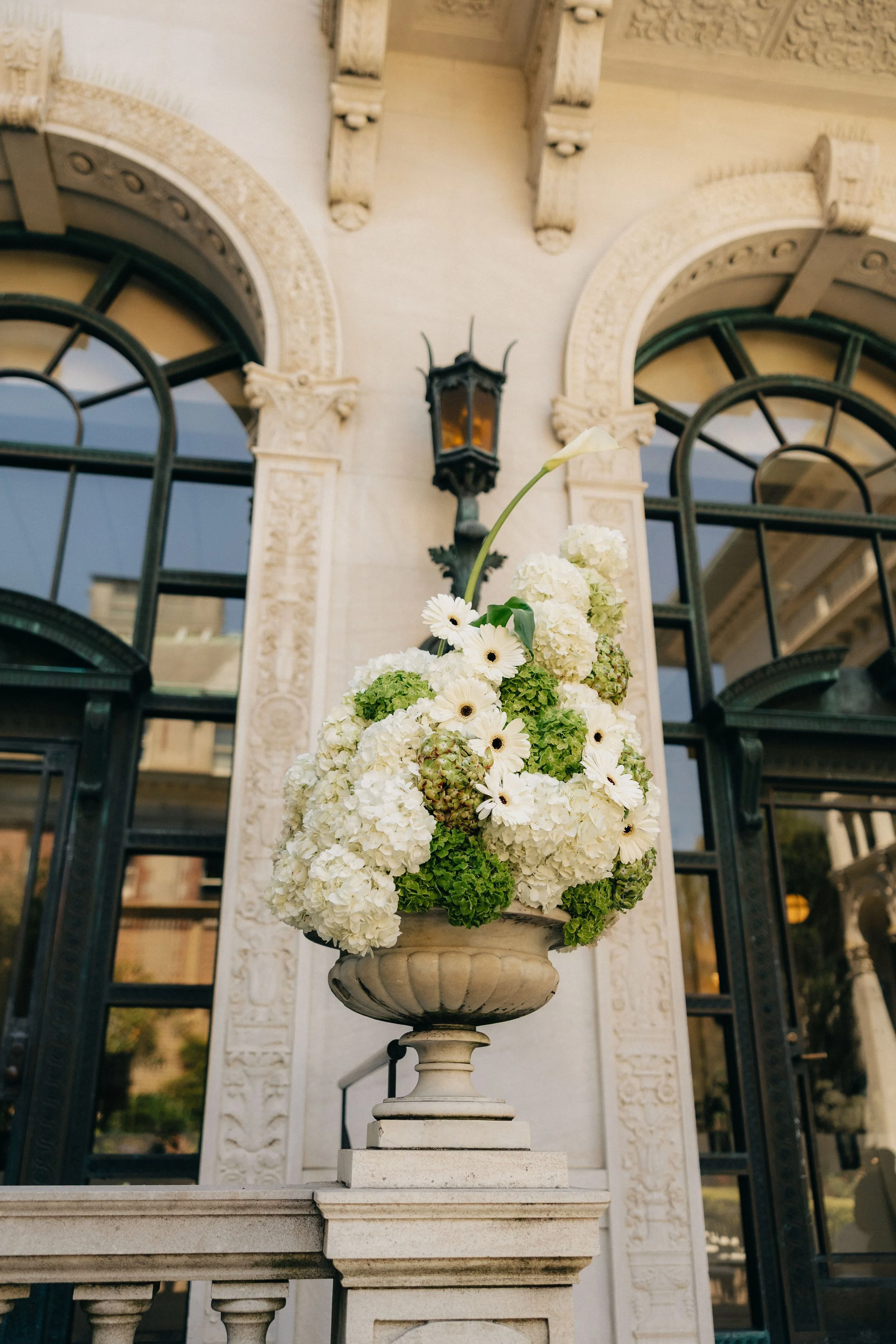 A decorative outdoor stone pedestal holding a bouquet of white flowers, including hydrangeas, daisies, and calla lilies, in front of an ornate building facade with arched windows and decorative architectural details.