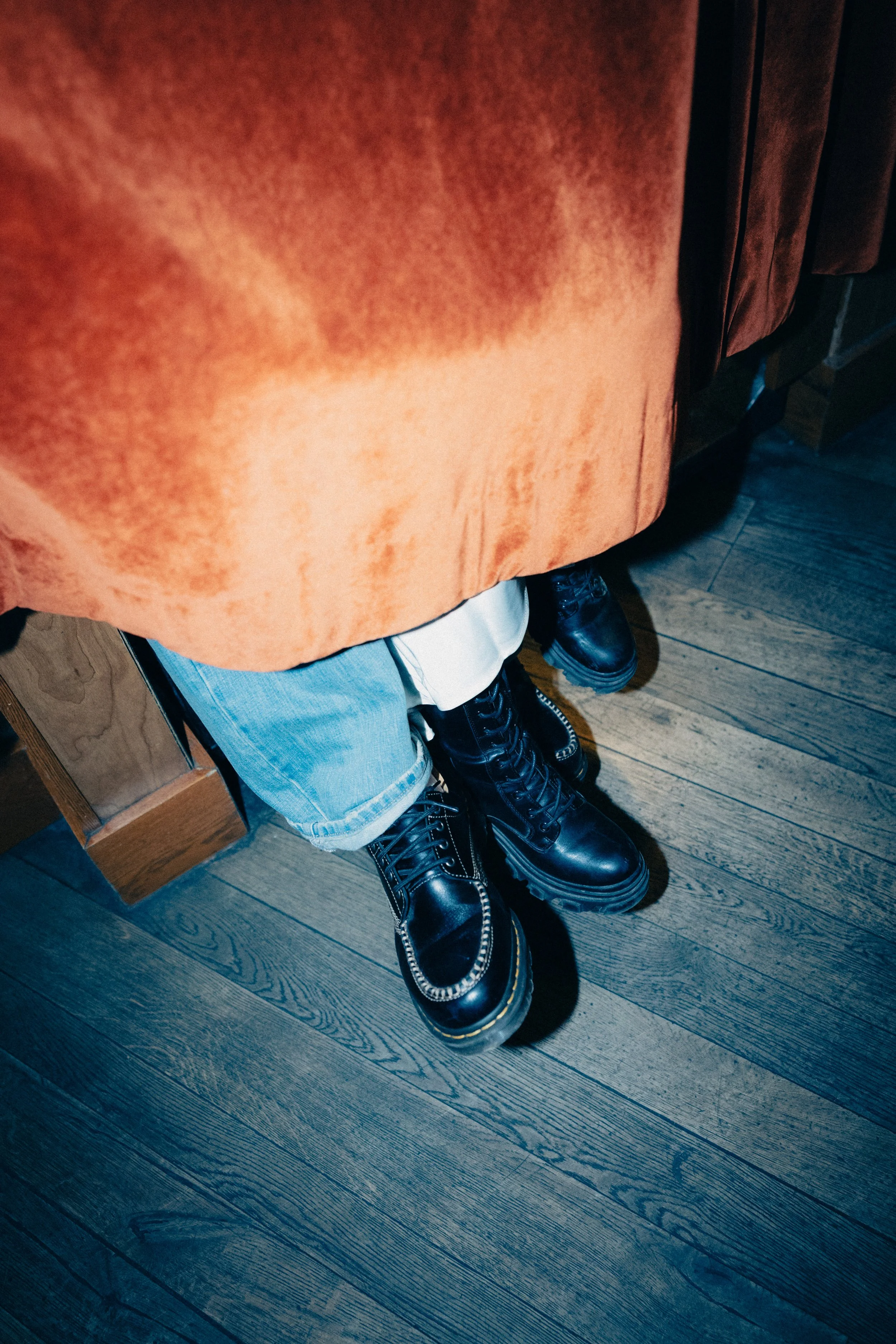 Person wearing black combat boots, light blue jeans with rolled cuffs, an orange jacket, and a white shirt underneath, standing on a wooden floor.