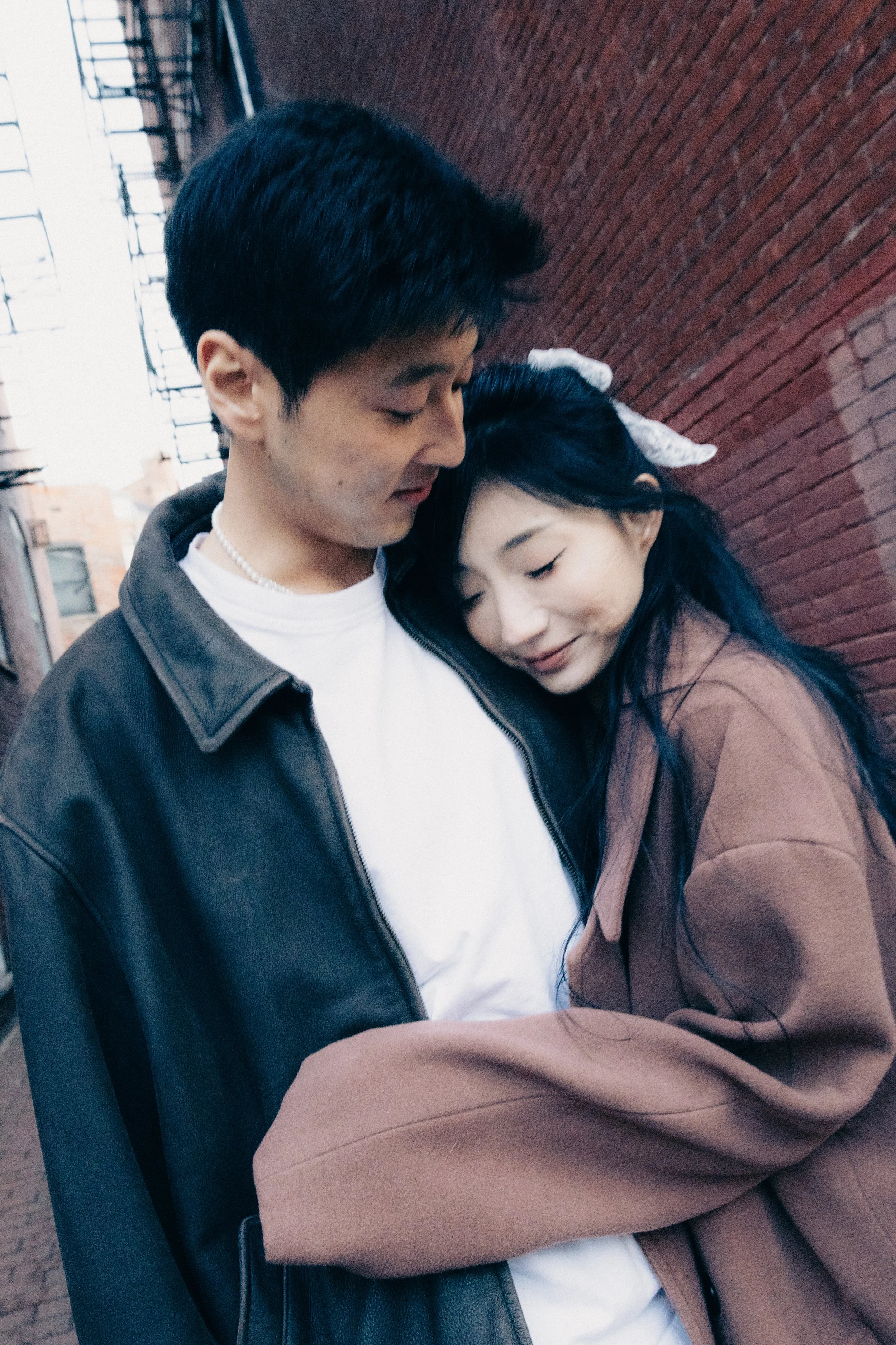 A young couple is sharing an intimate and affectionate moment outdoors against a red brick wall, with the woman resting her head on the man's chest.
