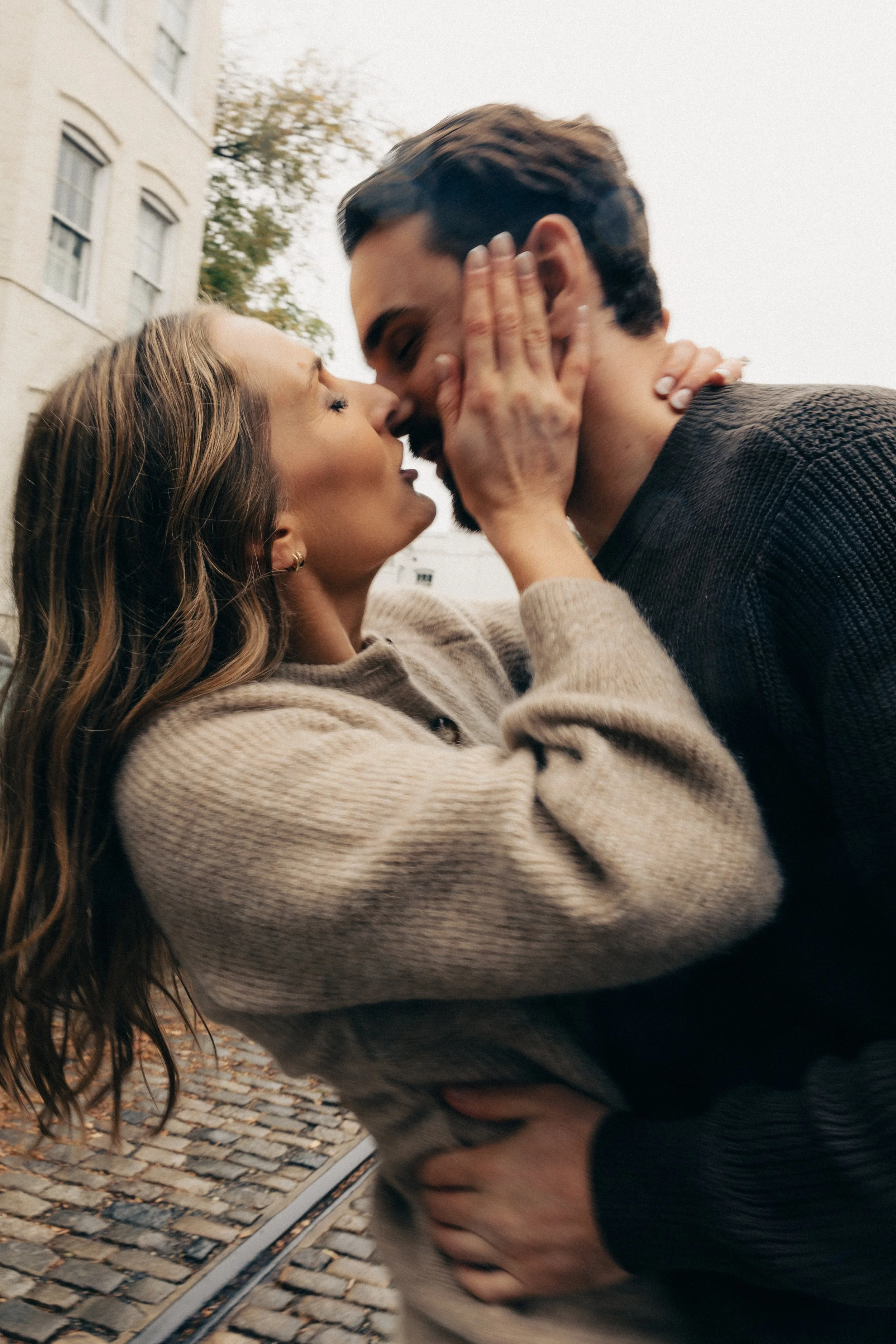 A couple is kissing outdoors, with the woman holding the man's face and the man gently holding the woman's waist. They are on a cobblestone street near buildings.
