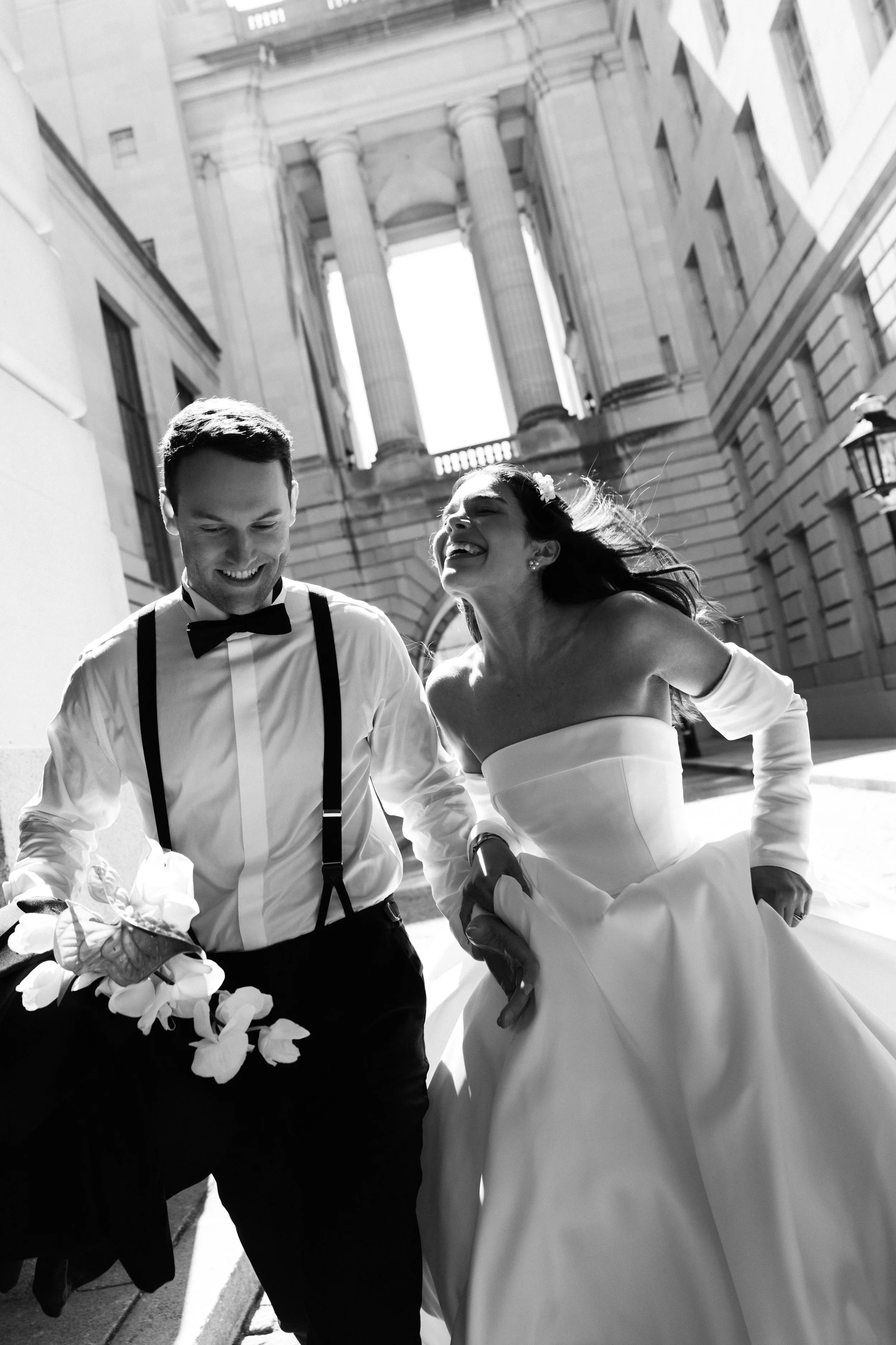 A black-and-white photo of a smiling couple, dressed in wedding attire, walking outdoors in front of large classical-style buildings with columns. The man is wearing a bow tie, suspenders, and holding a bouquet, while the woman is in a strapless wedding dress with a flower in her hair.