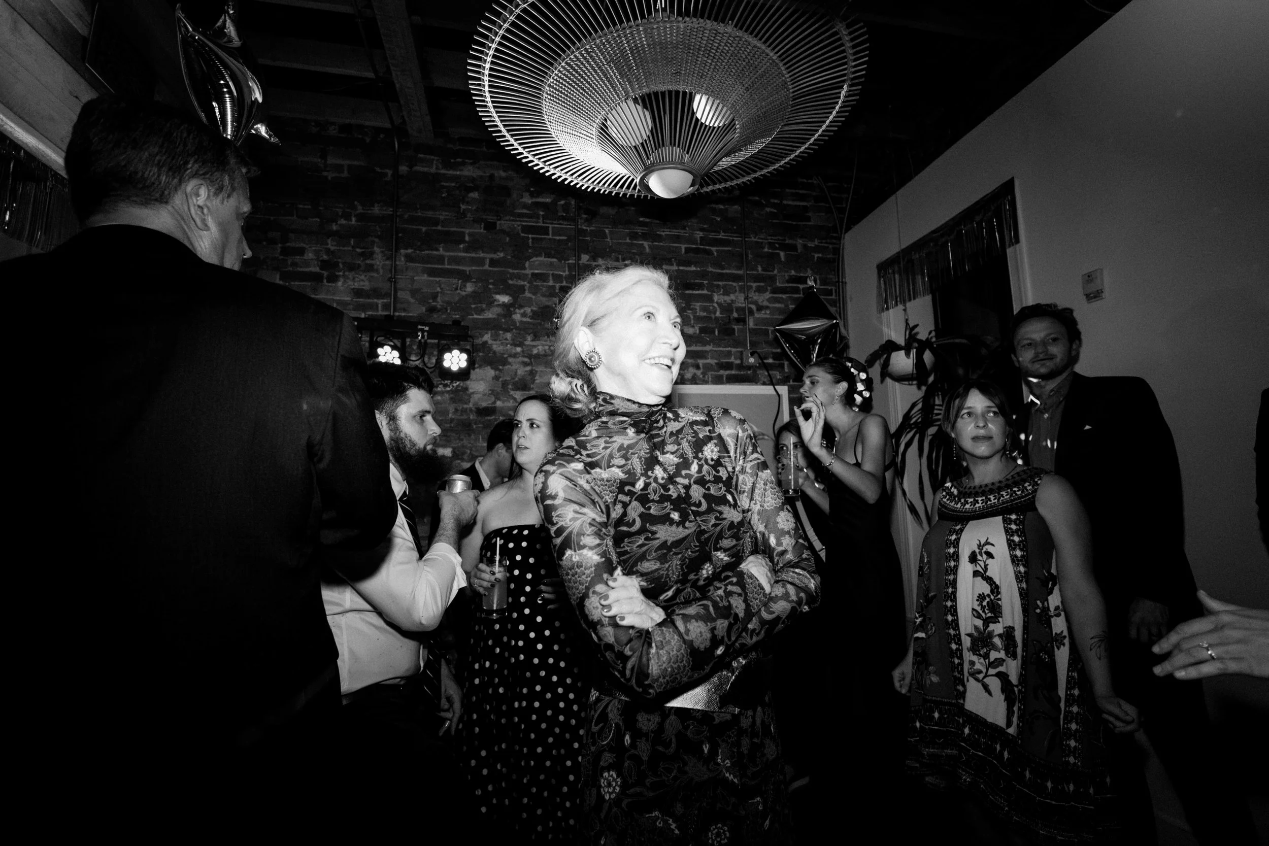 Group of people socializing at a party, with a woman smiling in the center, in a room with brick wall and modern light fixture
