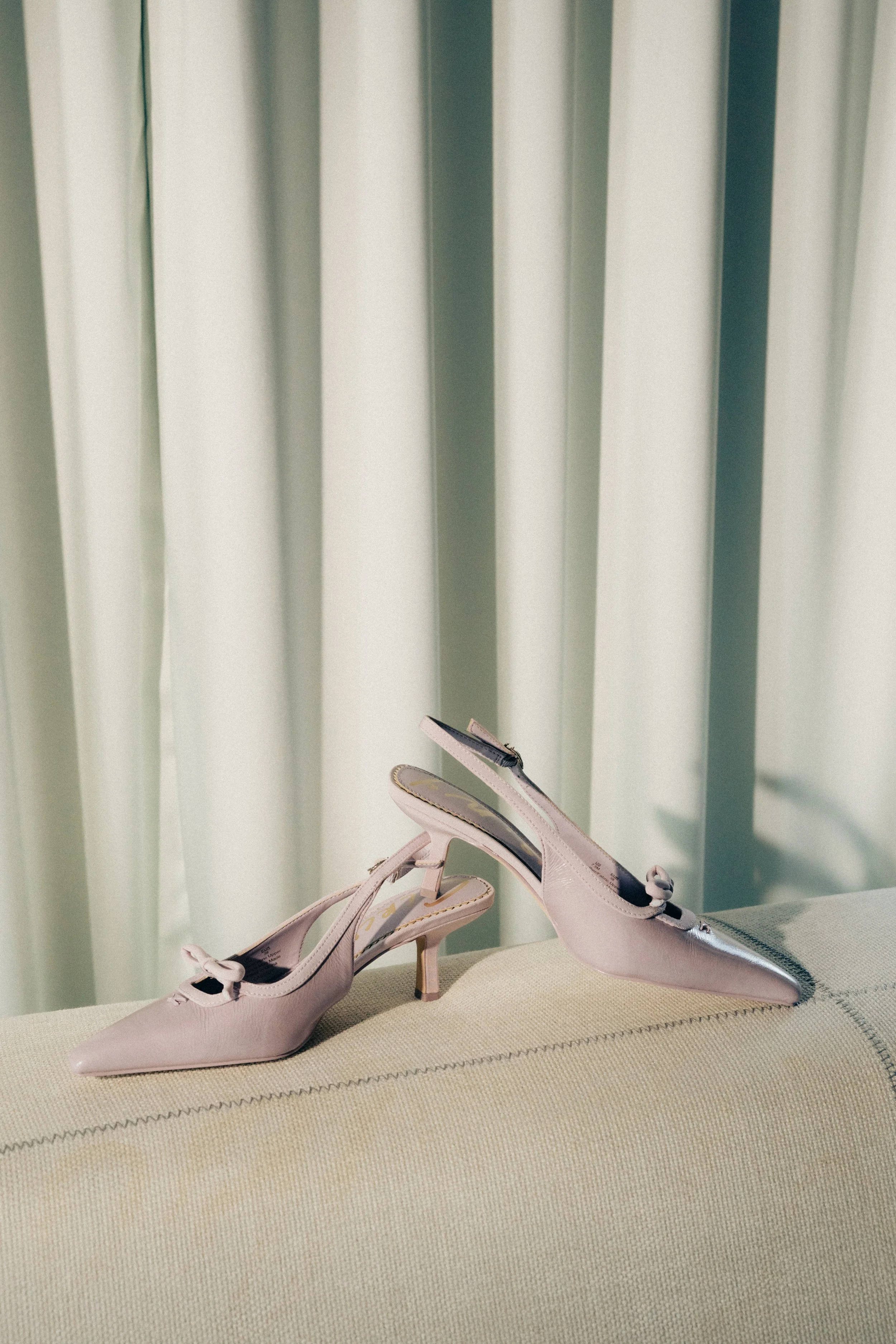 Pink high-heeled shoes with bows on a beige textured surface in front of green curtains.