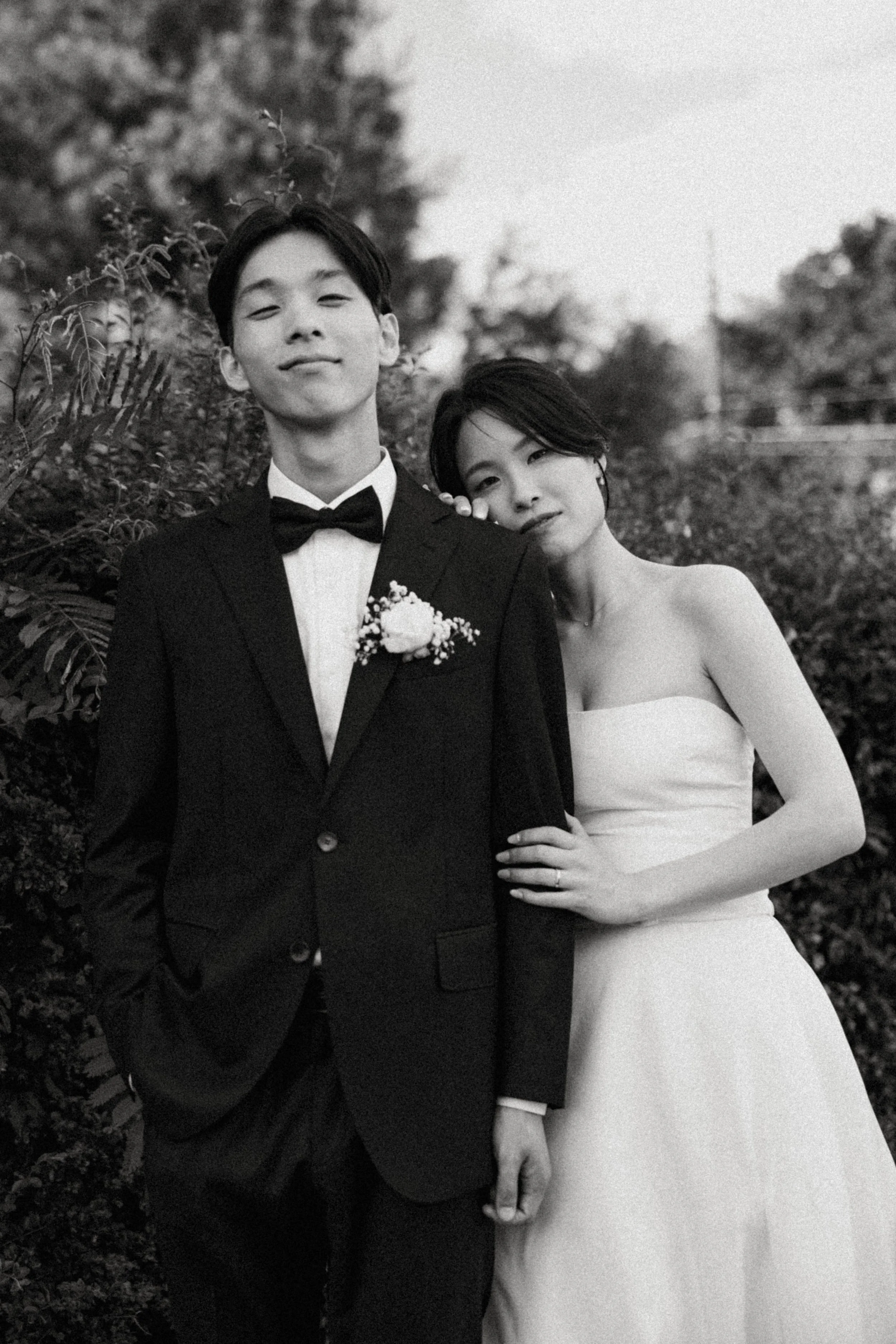 A black-and-white photo of a young man in a tuxedo with a flower boutonniere and a woman in a strapless dress, standing outdoors with trees and bushes in the background.