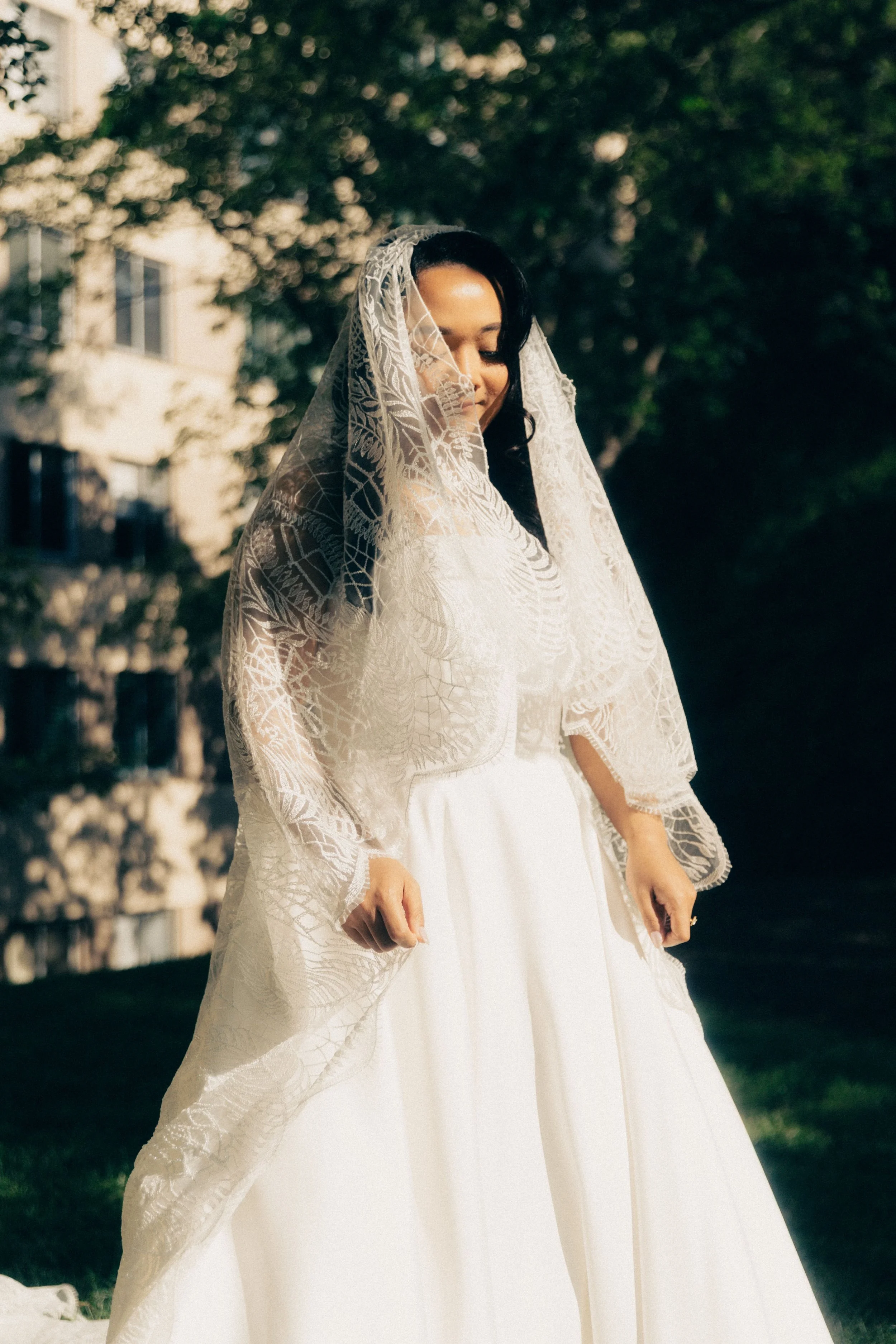 A woman wearing a white wedding dress and lace veil standing outdoors with trees and buildings in the background.