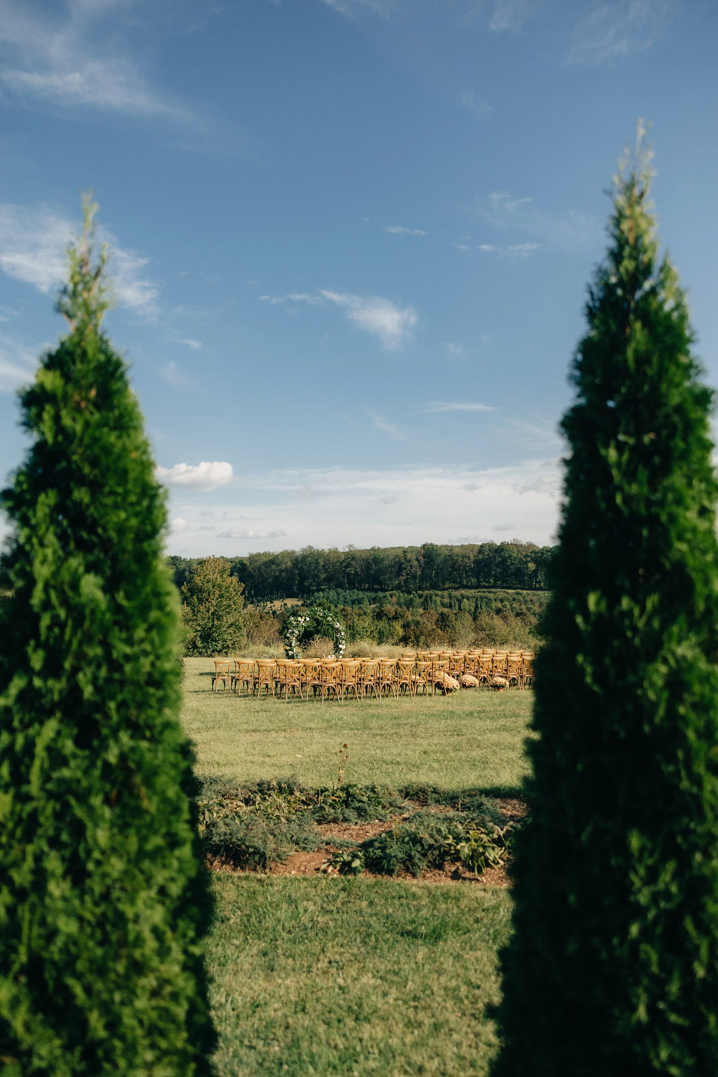 Outdoor wedding ceremony setup with wooden chairs arranged in rows, a floral arch at the front, under a clear blue sky with a few clouds, surrounded by green trees and landscape.