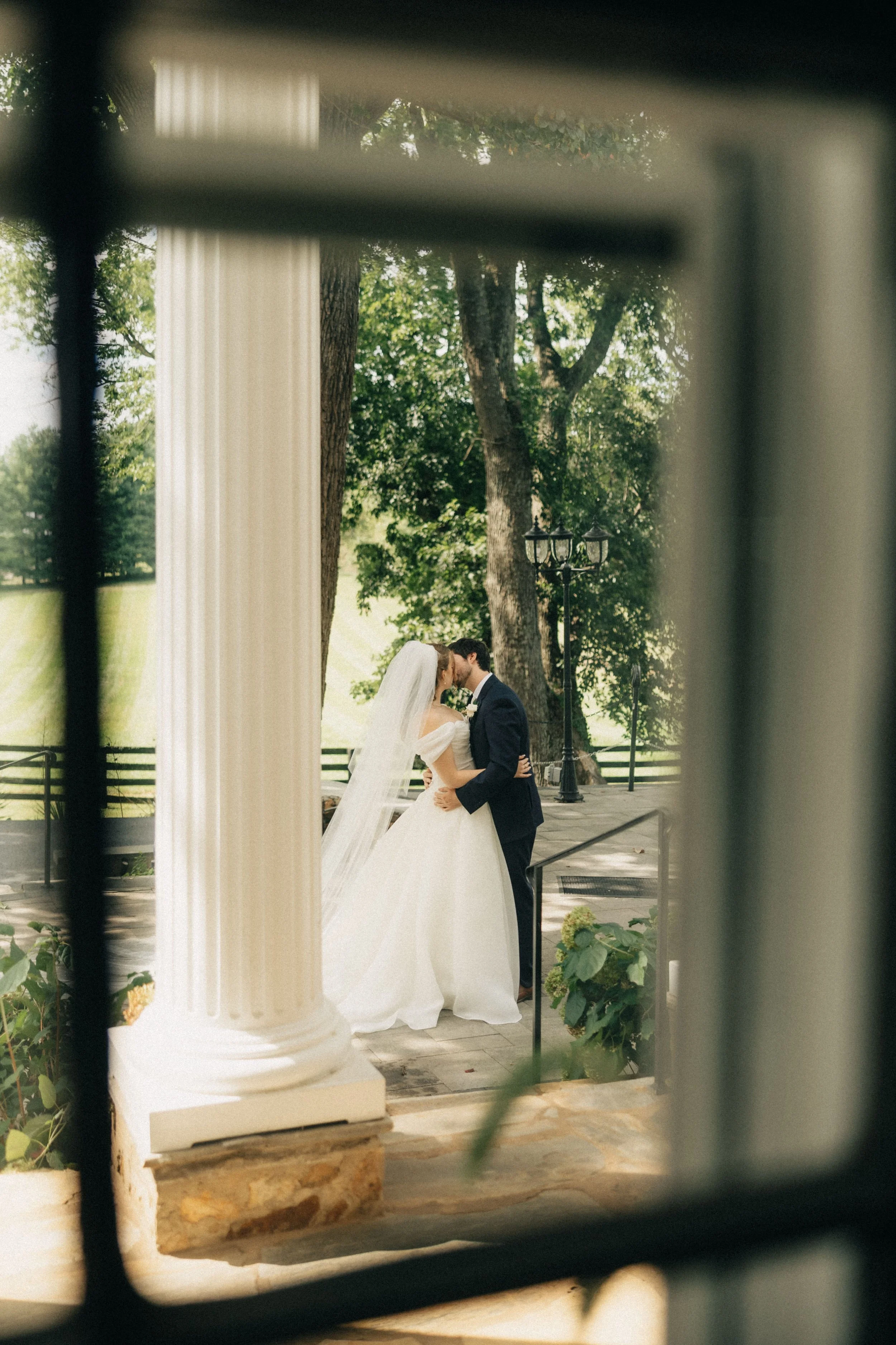 A bride and groom share a kiss outdoors, seen through black iron bars of a gate or fence. The bride wears a white wedding gown with a veil, and the groom wears a dark suit. The scene is set in a green park with trees and a street lamp in the background.