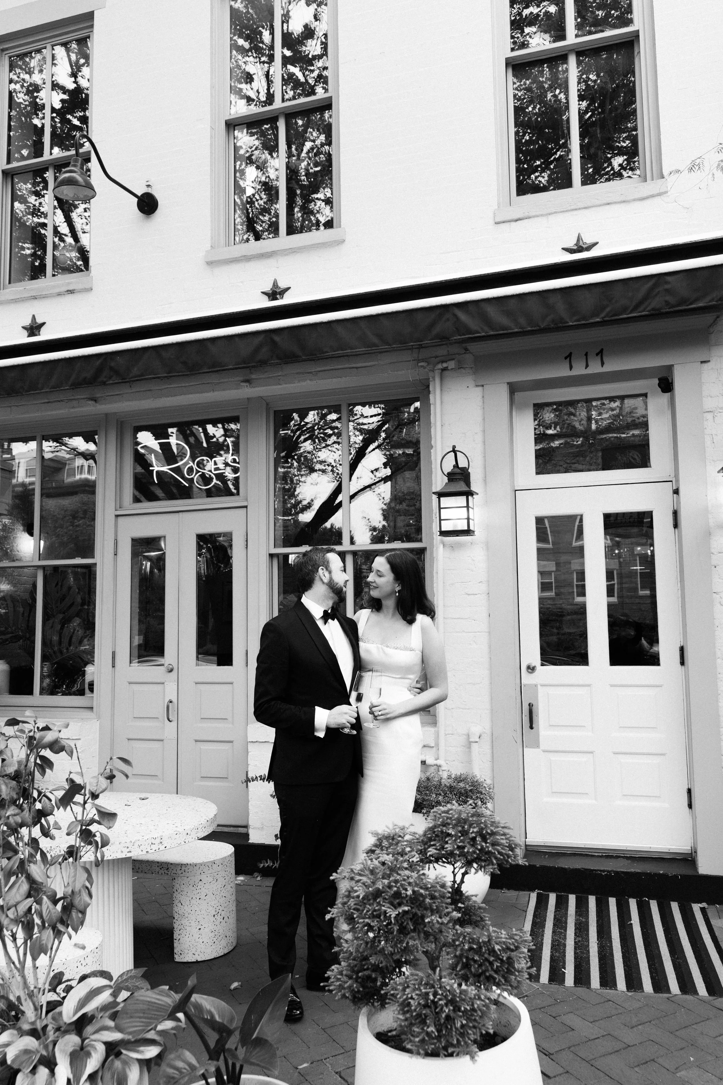 A black-and-white photo of a man in a tuxedo and a woman in an elegant dress standing outside a building with large windows and doors, holding glasses of wine and looking at each other. The building has a sign that says 'Roses' in the window.
