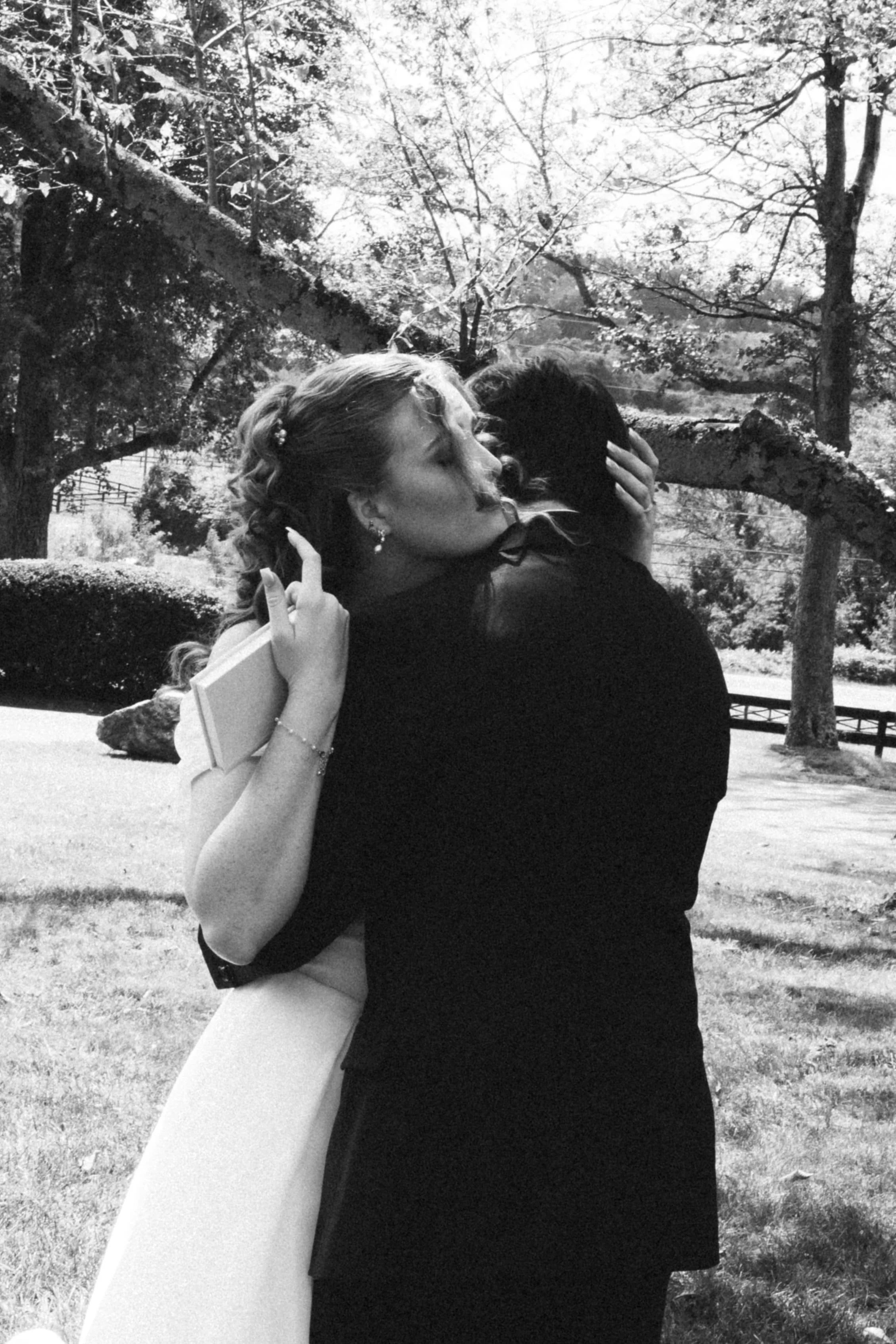 A black and white photo of a couple embracing outdoors in a park with trees and grass in the background. The woman is holding a book and appears to be giving a gentle kiss or whispering to the man.