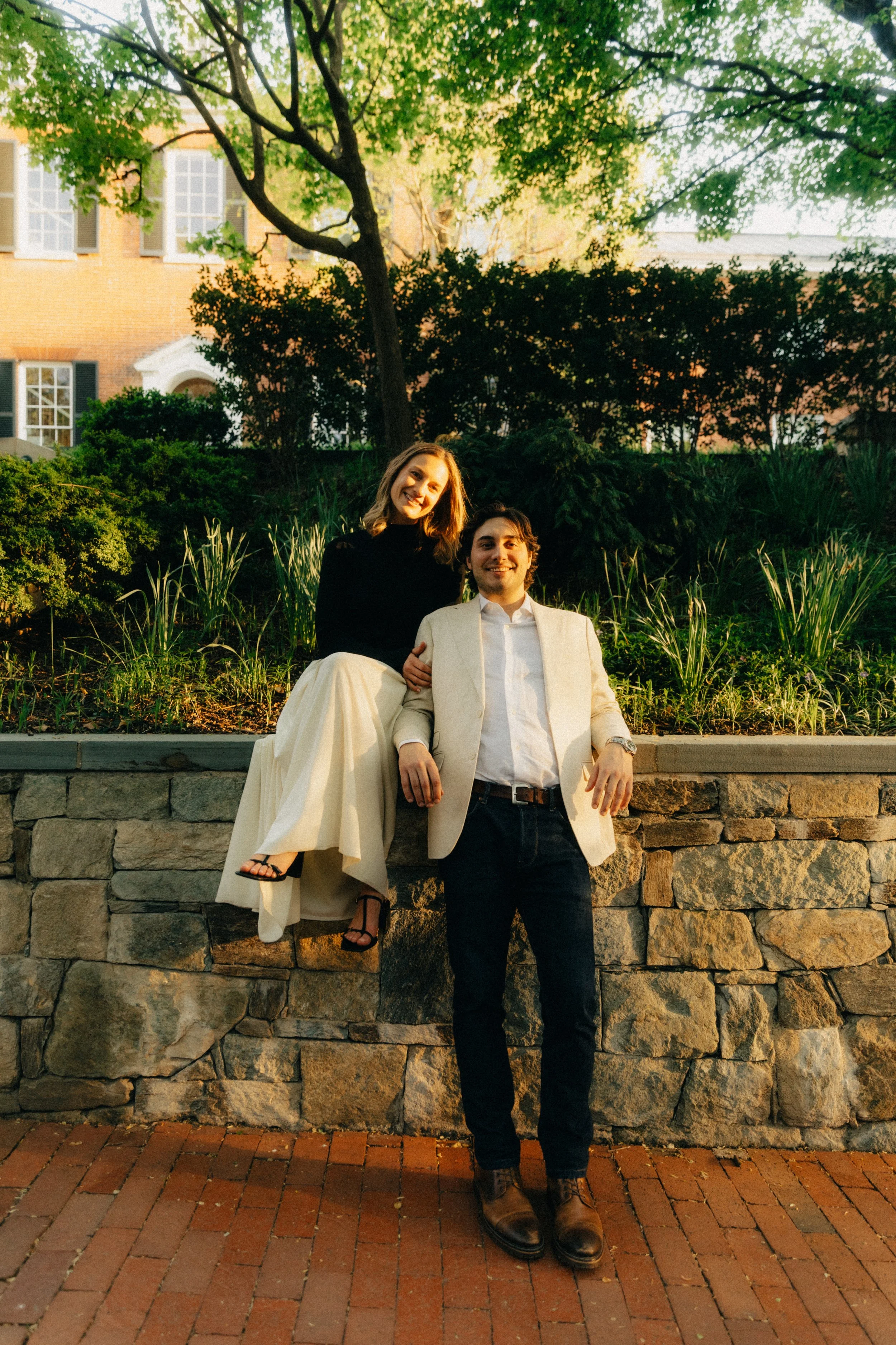 A man and woman posing outdoors at night, with the woman sitting on a stone wall and the man standing in front of it. The woman is smiling and wearing a black top and cream-colored pants, while the man is smiling and dressed in a light blazer, white shirt, and dark pants. They are surrounded by greenery and in front of a building with brick walls and large windows, illuminated by warm outdoor lighting.