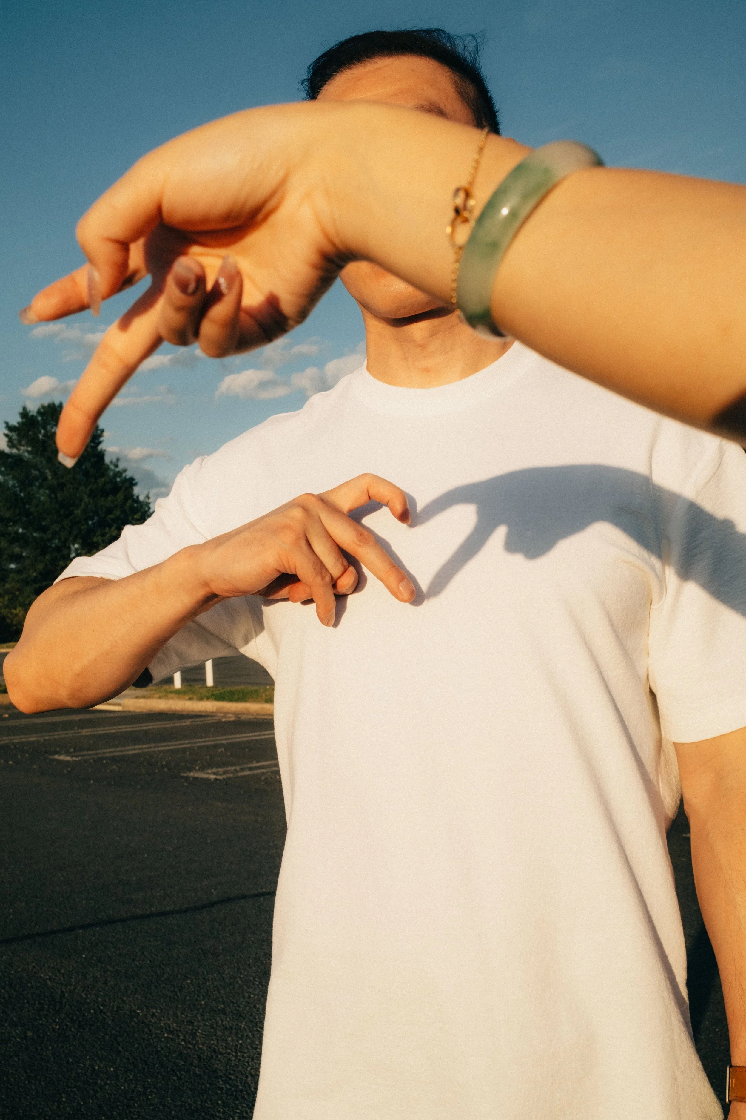 Person wearing a white shirt making a peace sign with both hands, standing outdoors during daytime with blue sky and trees in the background.