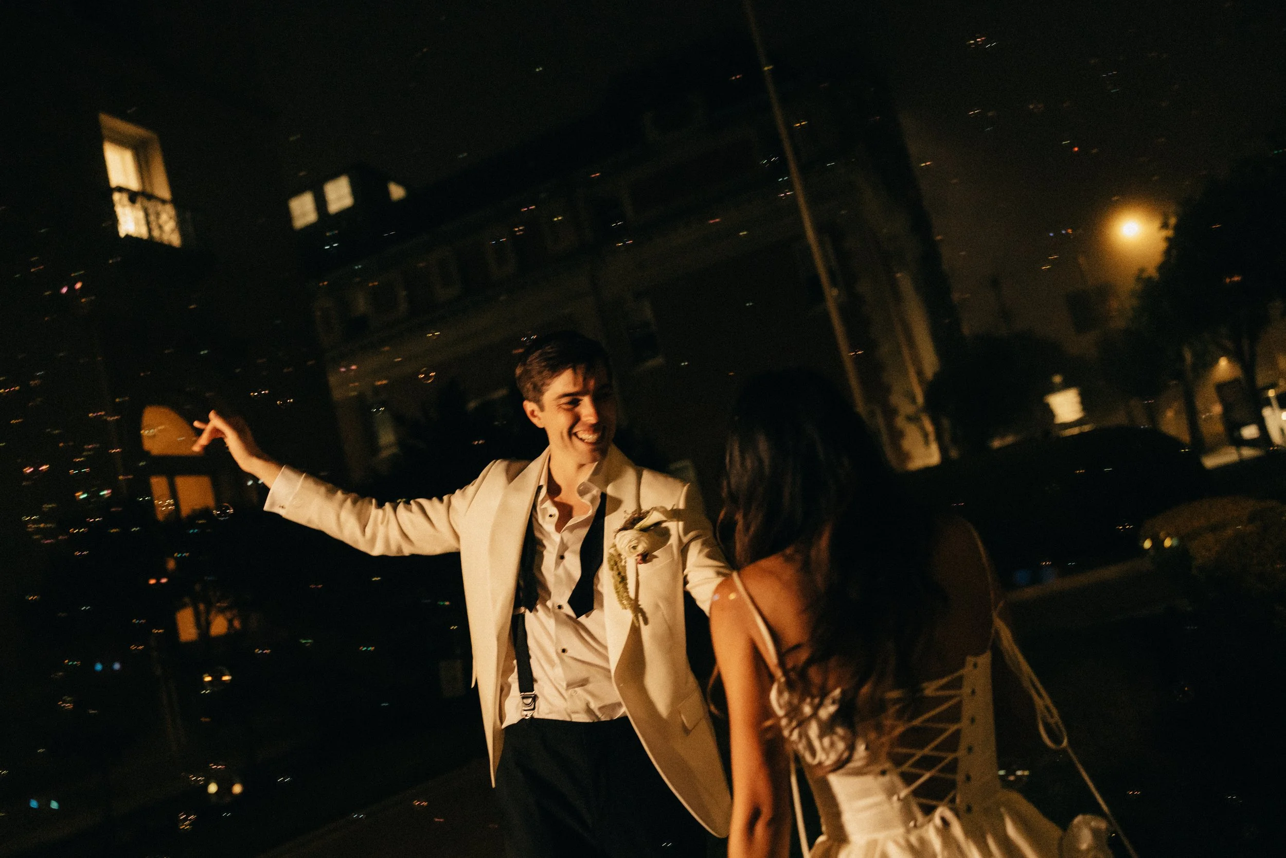 A young man dressed in a beige suit with a black tie, smiling, dancing, or celebrating with an unseen woman during nighttime outdoors with city lights in the background.