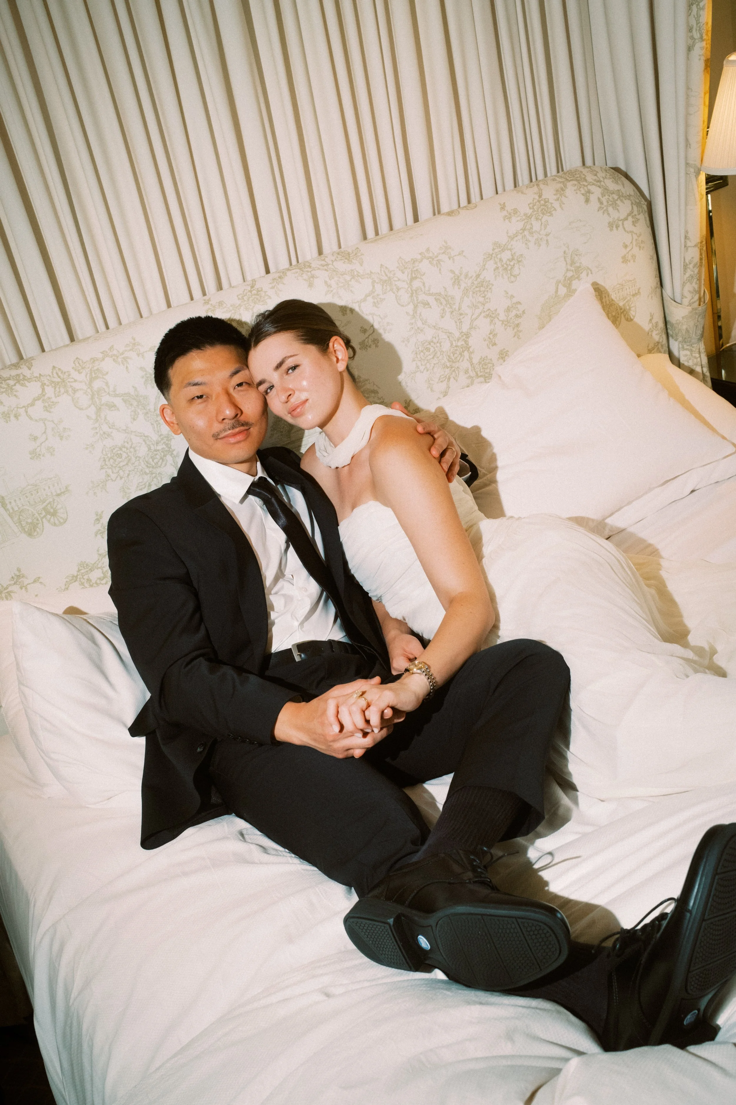 A man and a woman sitting close together on a hotel bed, smiling and holding hands. The man is wearing a black suit with a tie, and the woman is wearing a white dress. There is a headboard with floral upholstery, lamps on bedside tables, and curtains behind them.