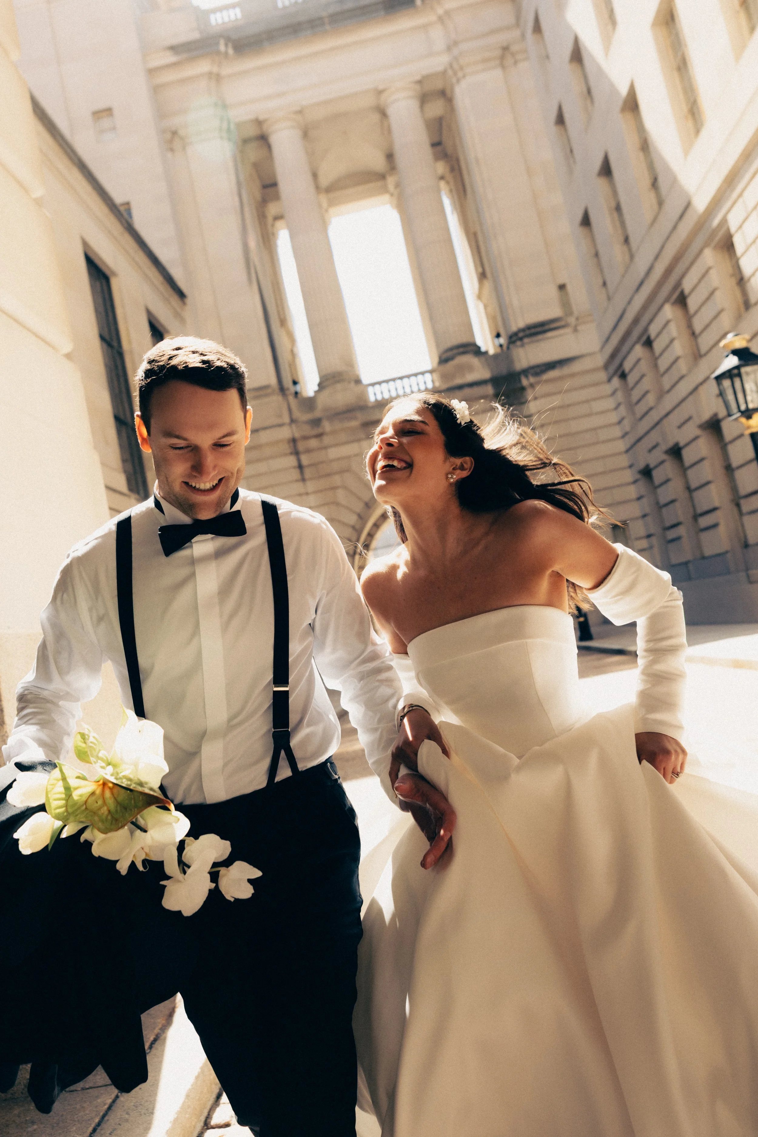 A smiling newlywed couple walking outside, with a grand historic building in the background. The groom is wearing a white shirt with suspenders and a bow tie, holding a bouquet of flowers. The bride is in a white wedding dress with an off-shoulder neckline, her hair flowing freely, and appears joyful.