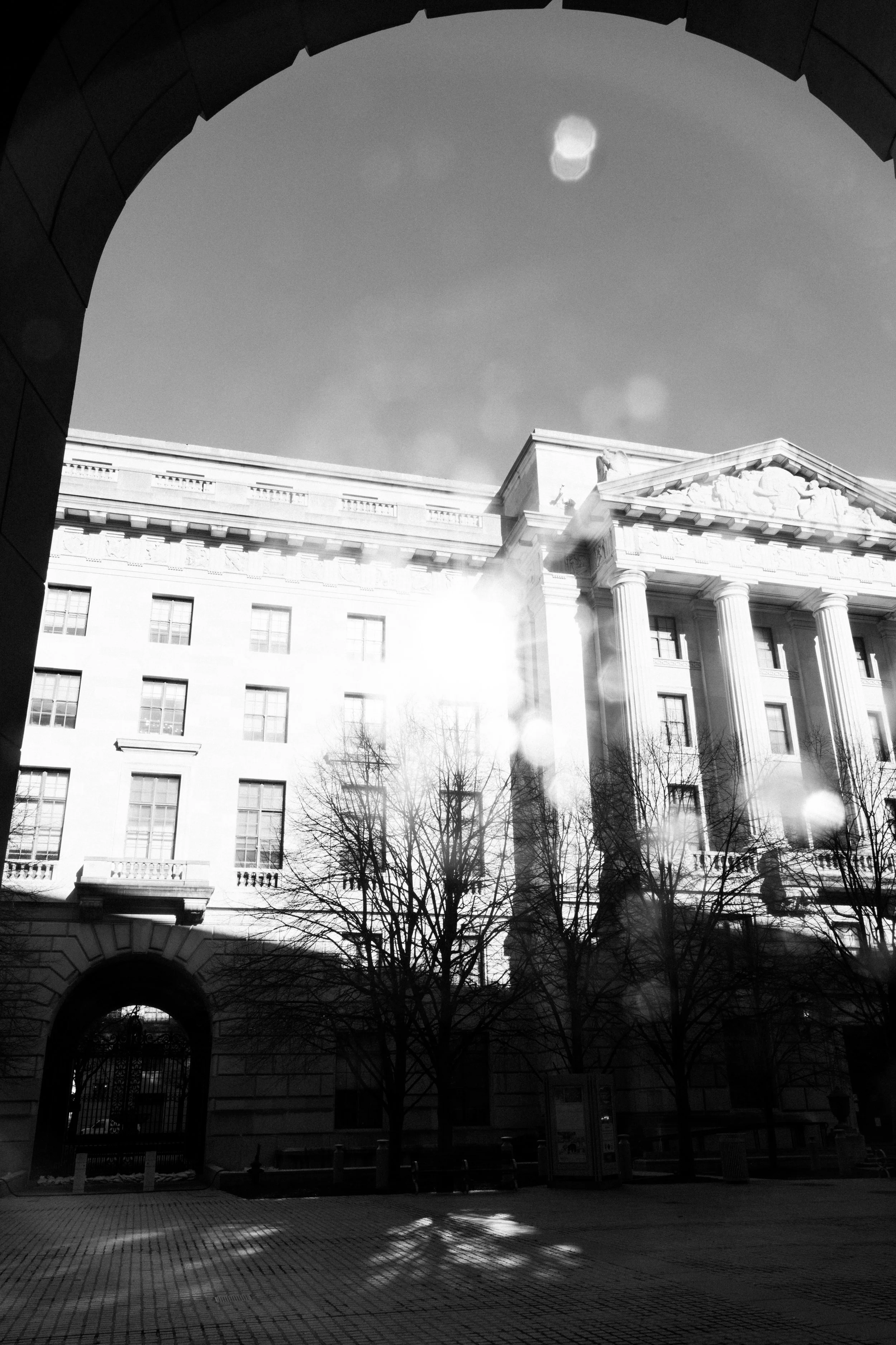 Black and white photo of a classical style building with columns and sculptural details, framed by an archway in the foreground, leafless trees, and a paved open space in front.