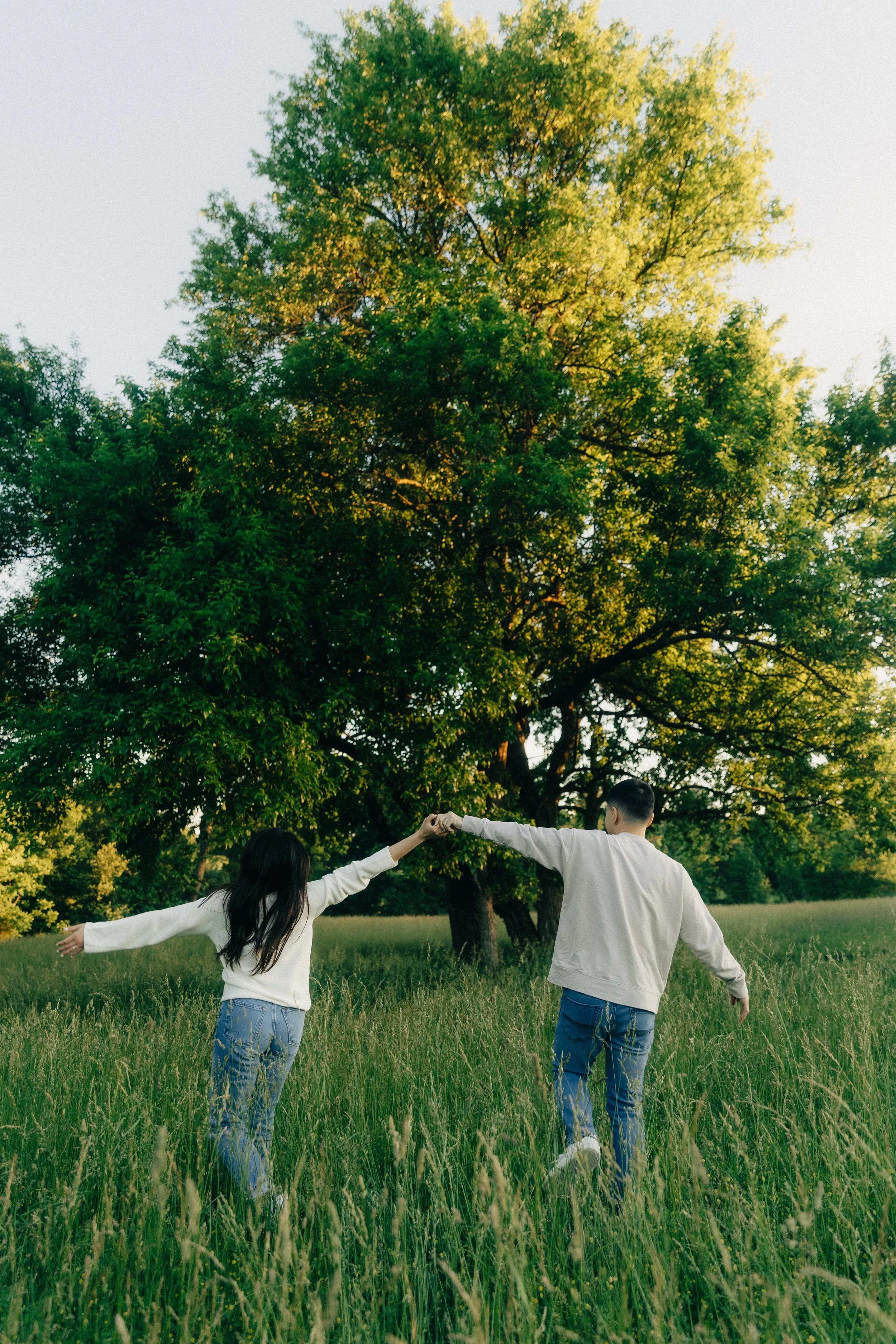 A young man and woman holding hands while walking through a grassy field with tall grass, trees, and clear sky in the background.