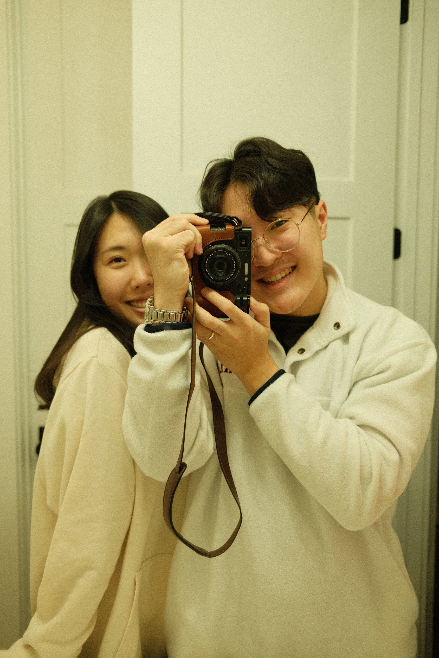 Two friends taking a mirror selfie, with one holding a camera and the other smiling, both wearing beige and cream-colored clothing.
