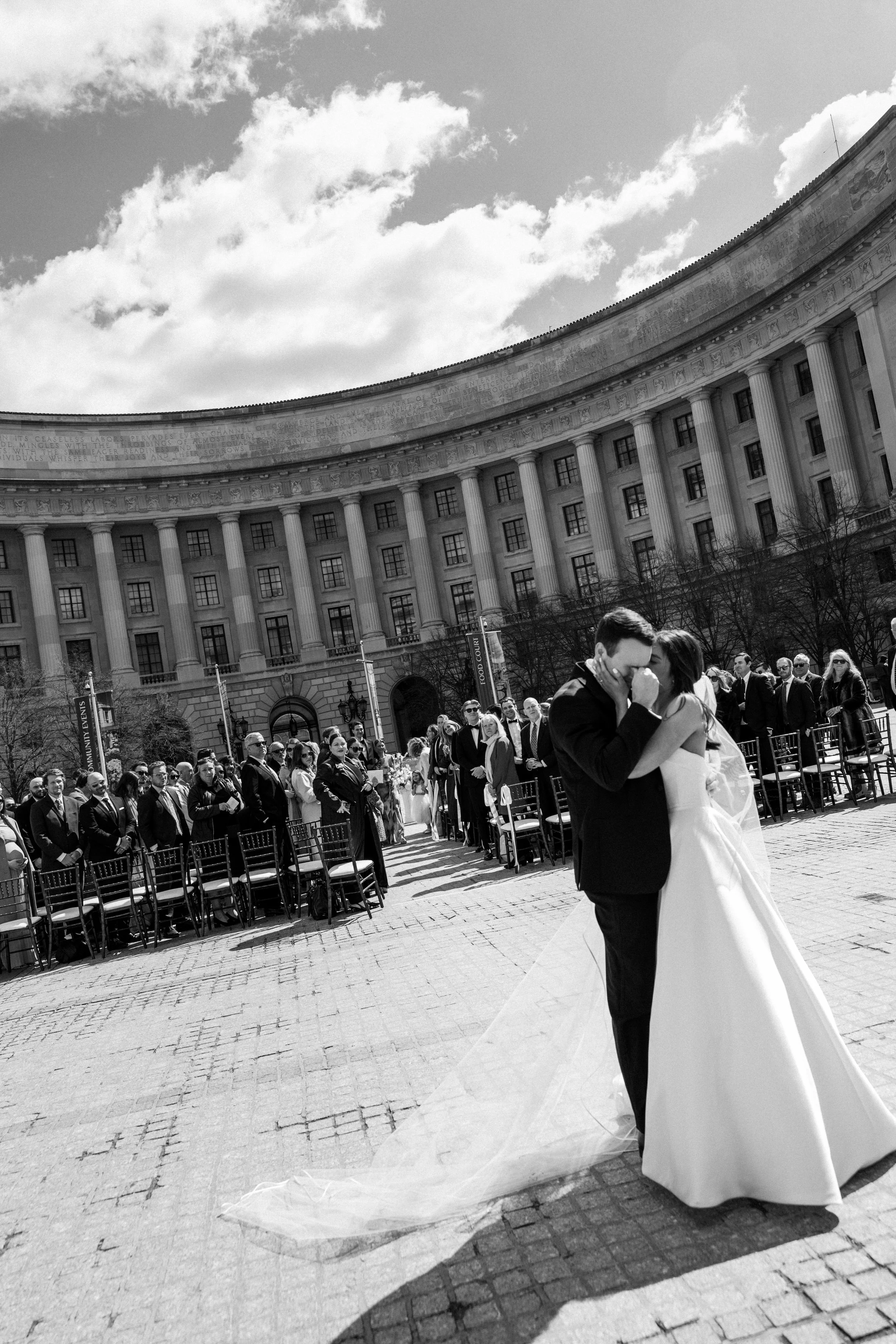 A couple dressed in wedding attire sharing a kiss in front of a crowd of people outside a large classical building, with a partly cloudy sky overhead.