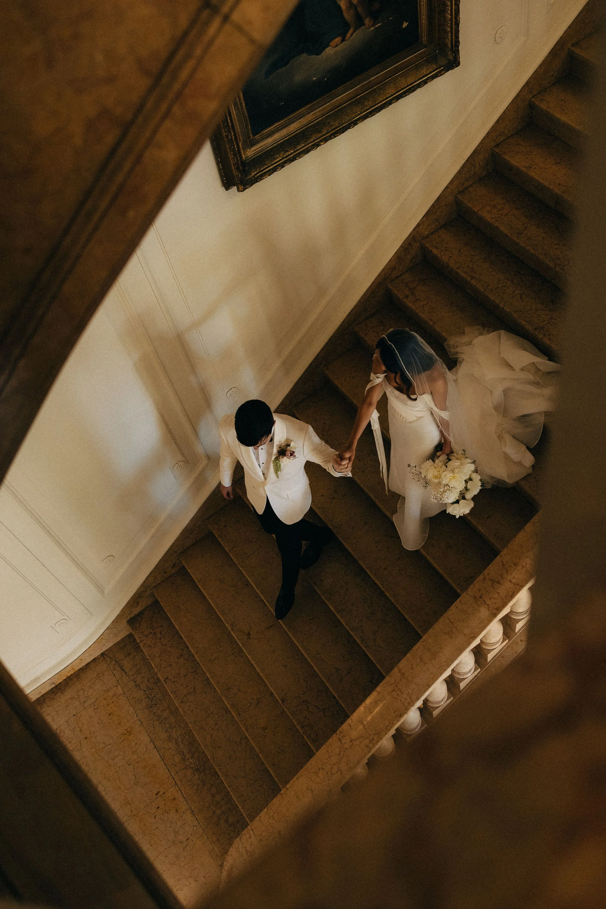 A bride and groom holding hands on a staircase, the bride in a white wedding dress with a bouquet, and the groom in a white suit, captured from above.