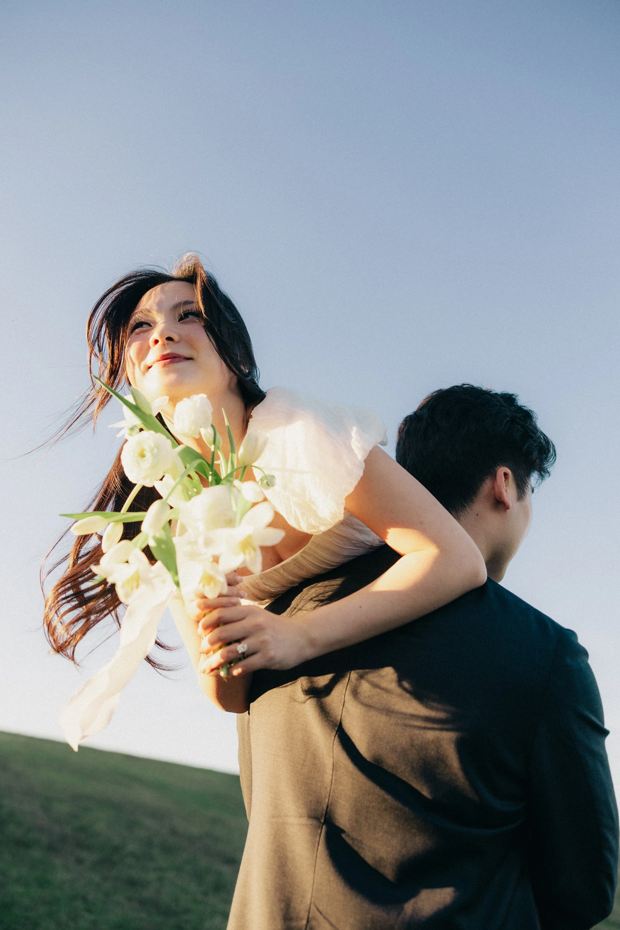 A woman with long dark hair holding a bouquet of white flowers with a man in a black jacket carrying her on his back outside against a clear blue sky.