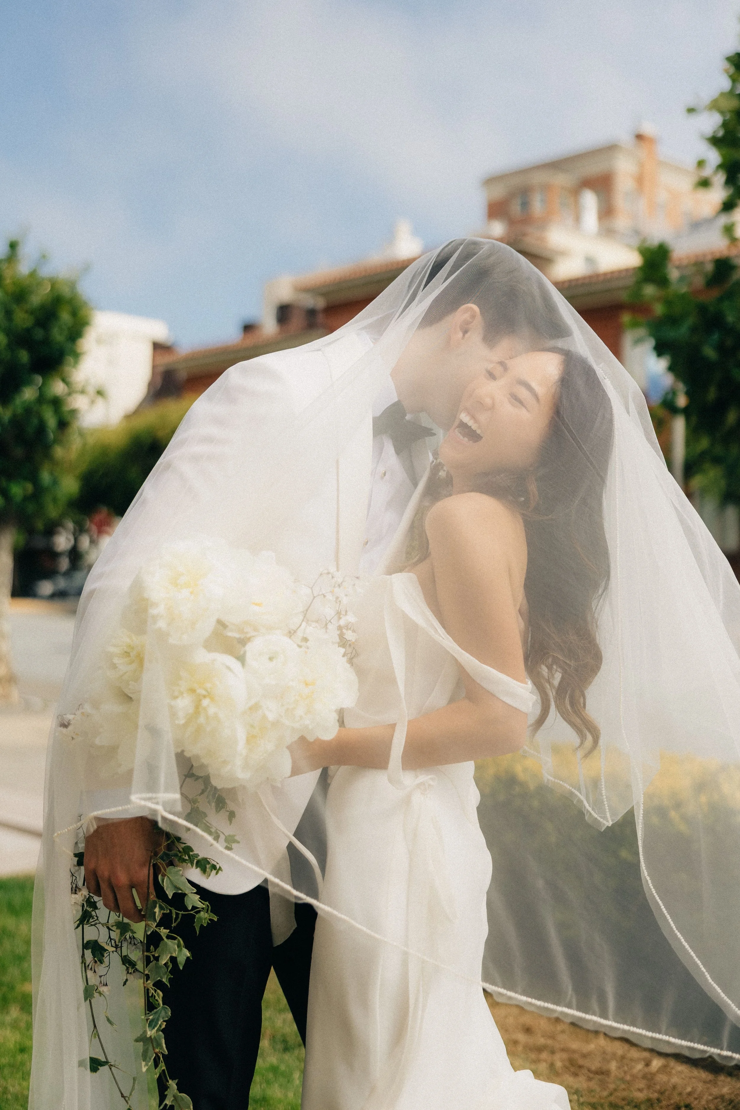 A bride and groom sharing a kiss under her veil outdoors on a sunny day.