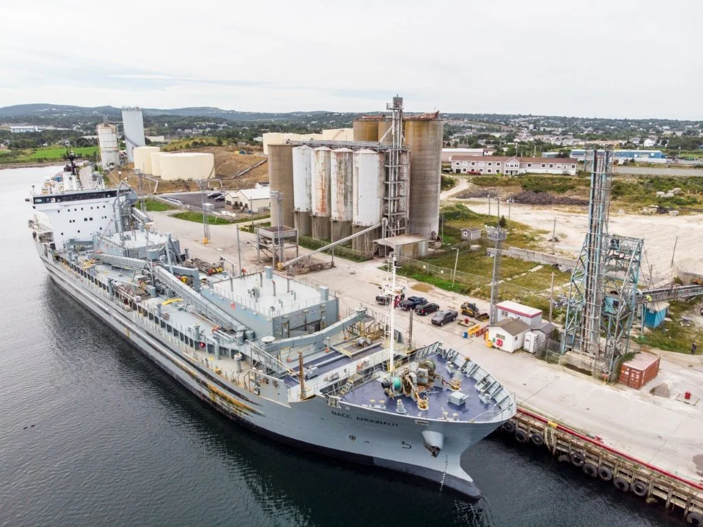 A large military ship docked at a port near industrial storage tanks and equipment, with a parking lot and buildings in the background.