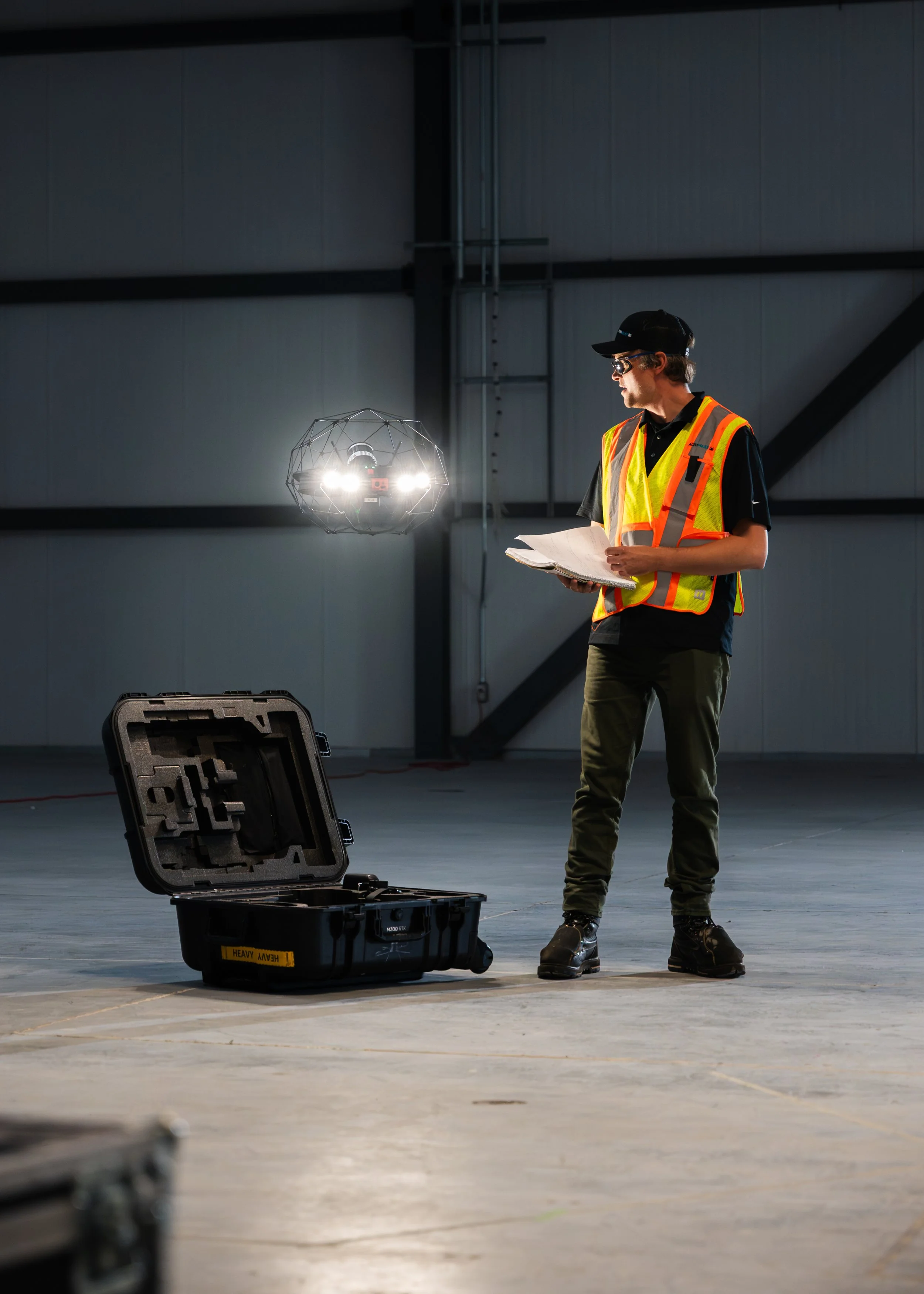 A man in a yellow safety vest and black cap holding papers stands inside an industrial hangar or warehouse, near a open black hard case labeled 'HEAVY' on the floor. There is a drone with lights hovering in the background, and the environment has a dark, high-ceiling, metallic wall.