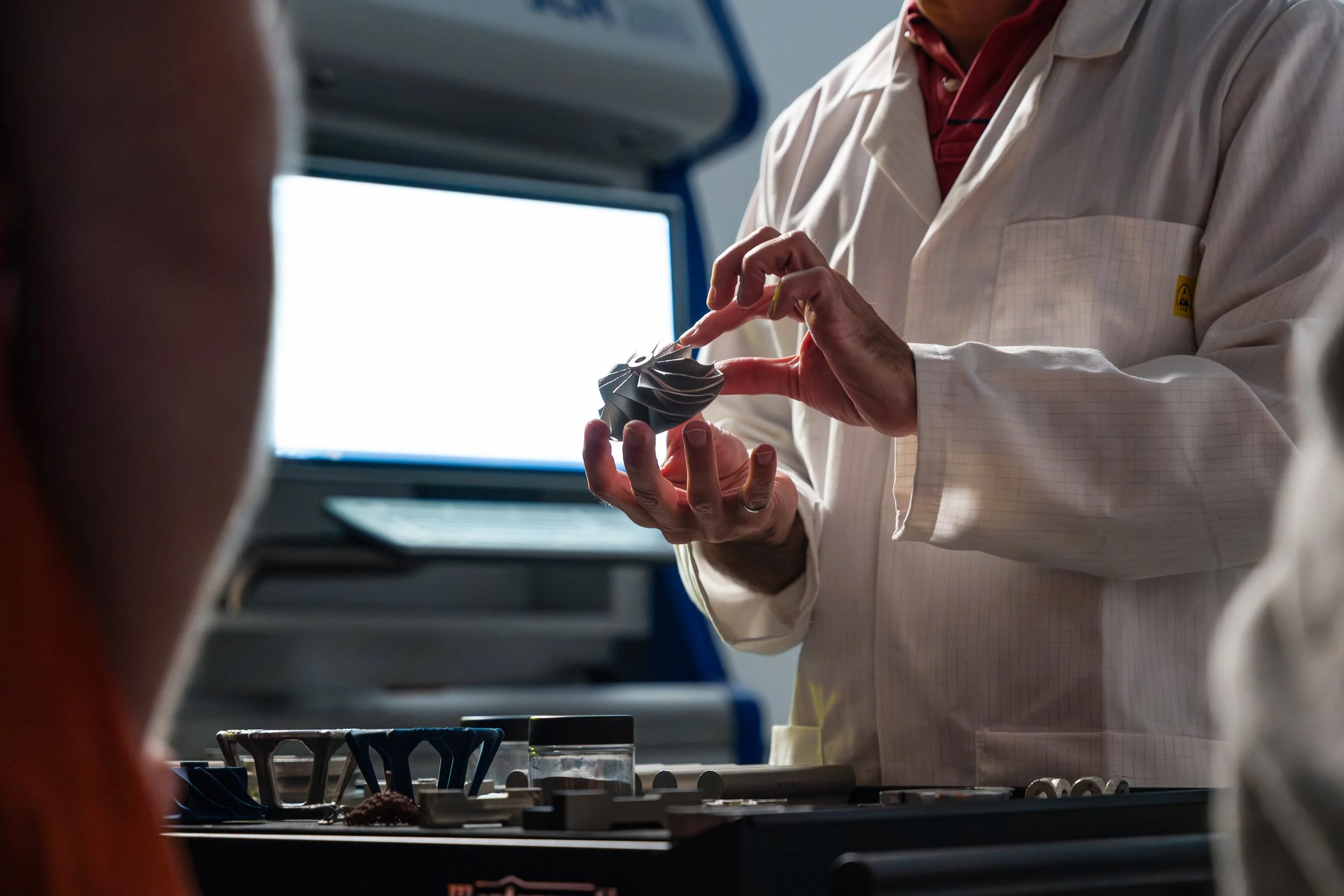 A person wearing a white lab coat and a red shirt is holding a small object, possibly a model or a component, while working at a table with tools and glassware. The person is blurred in the foreground, with a bright computer screen in the background.