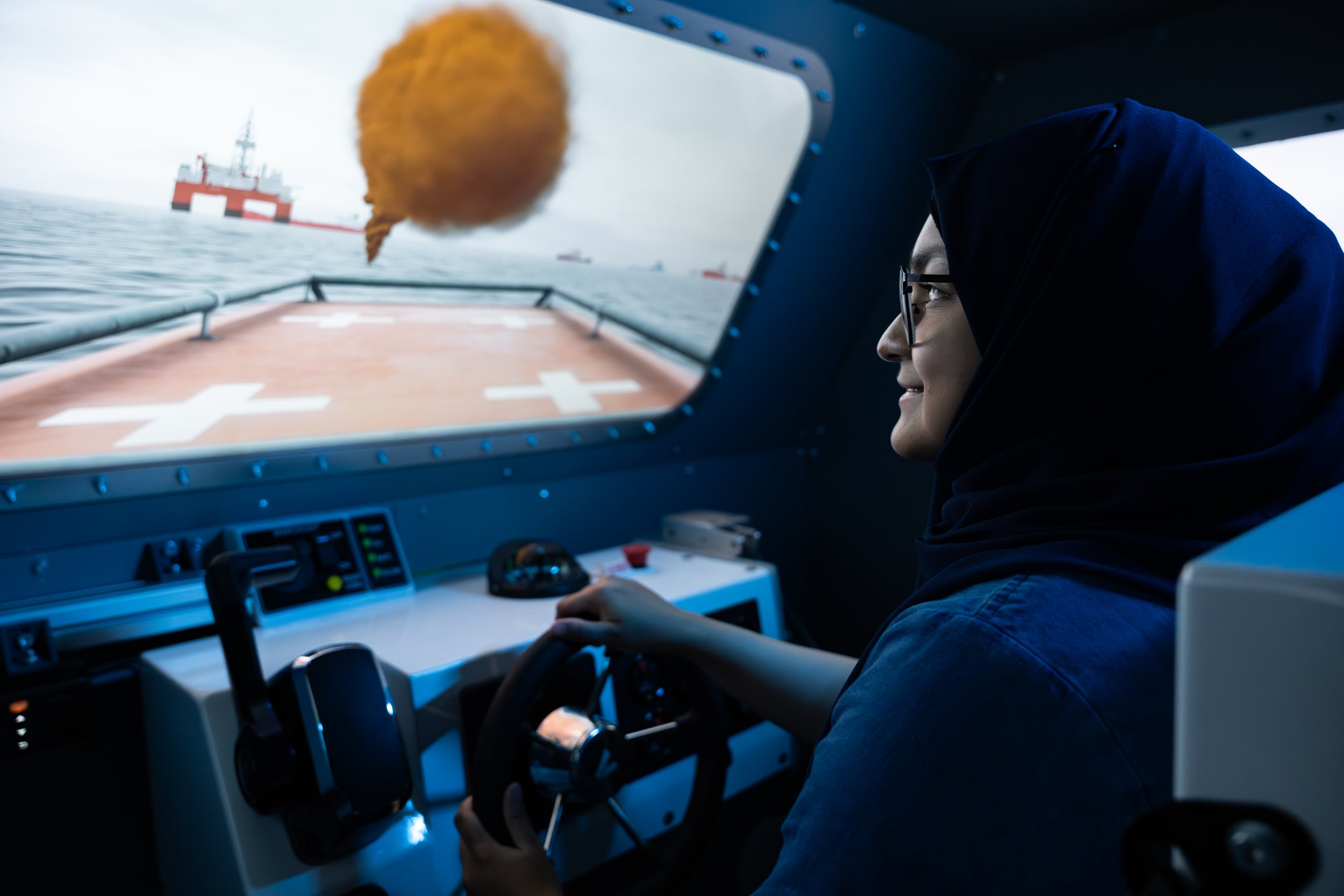 A person is using simulation technology training to navigate a ship, looking at a screen that shows the view from the ship, a small window at an offshore oil platform in the distance during cloudy weather.