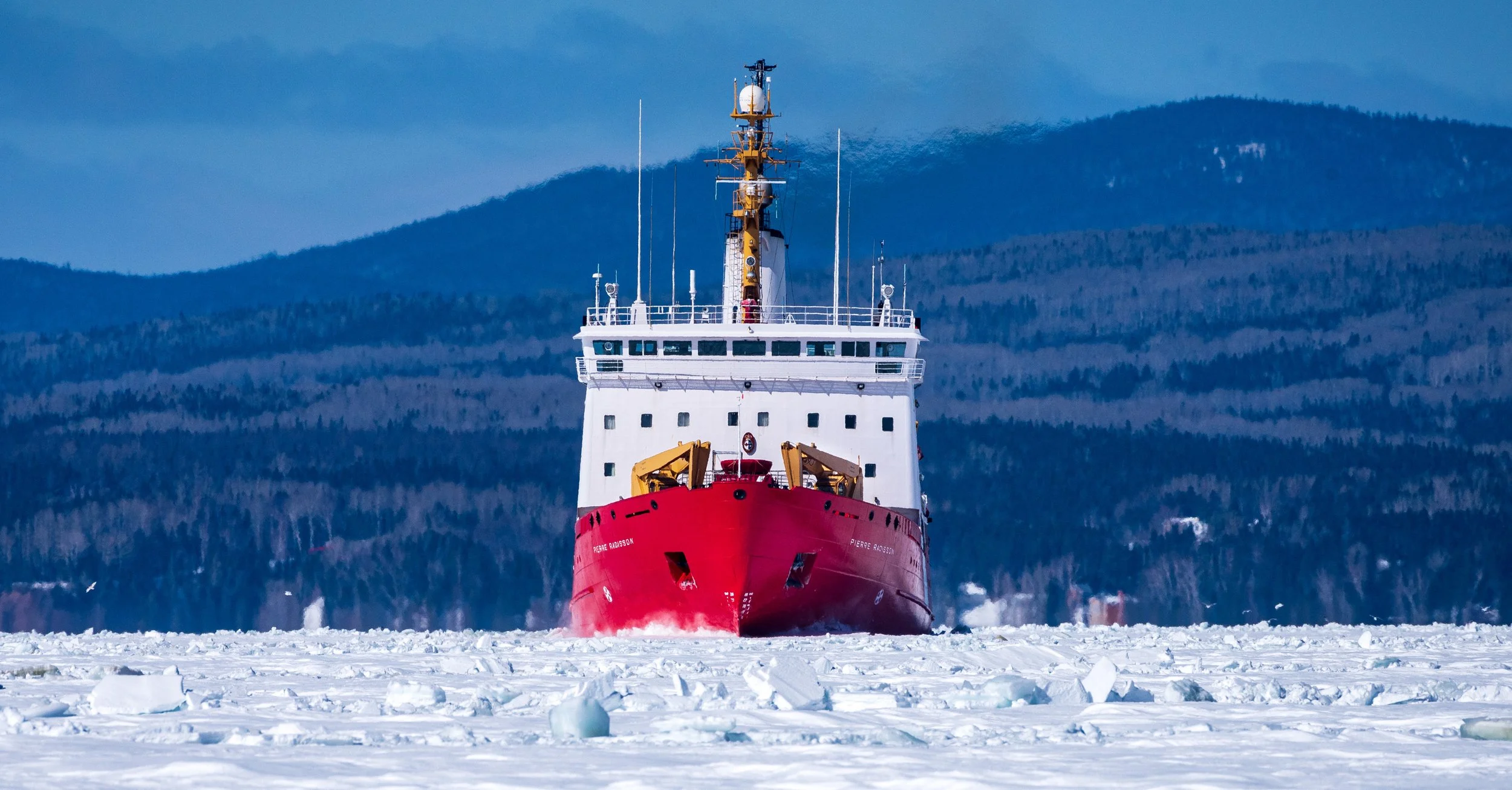 A large red ship navigating through icy waters with snow-covered mountains in the background.