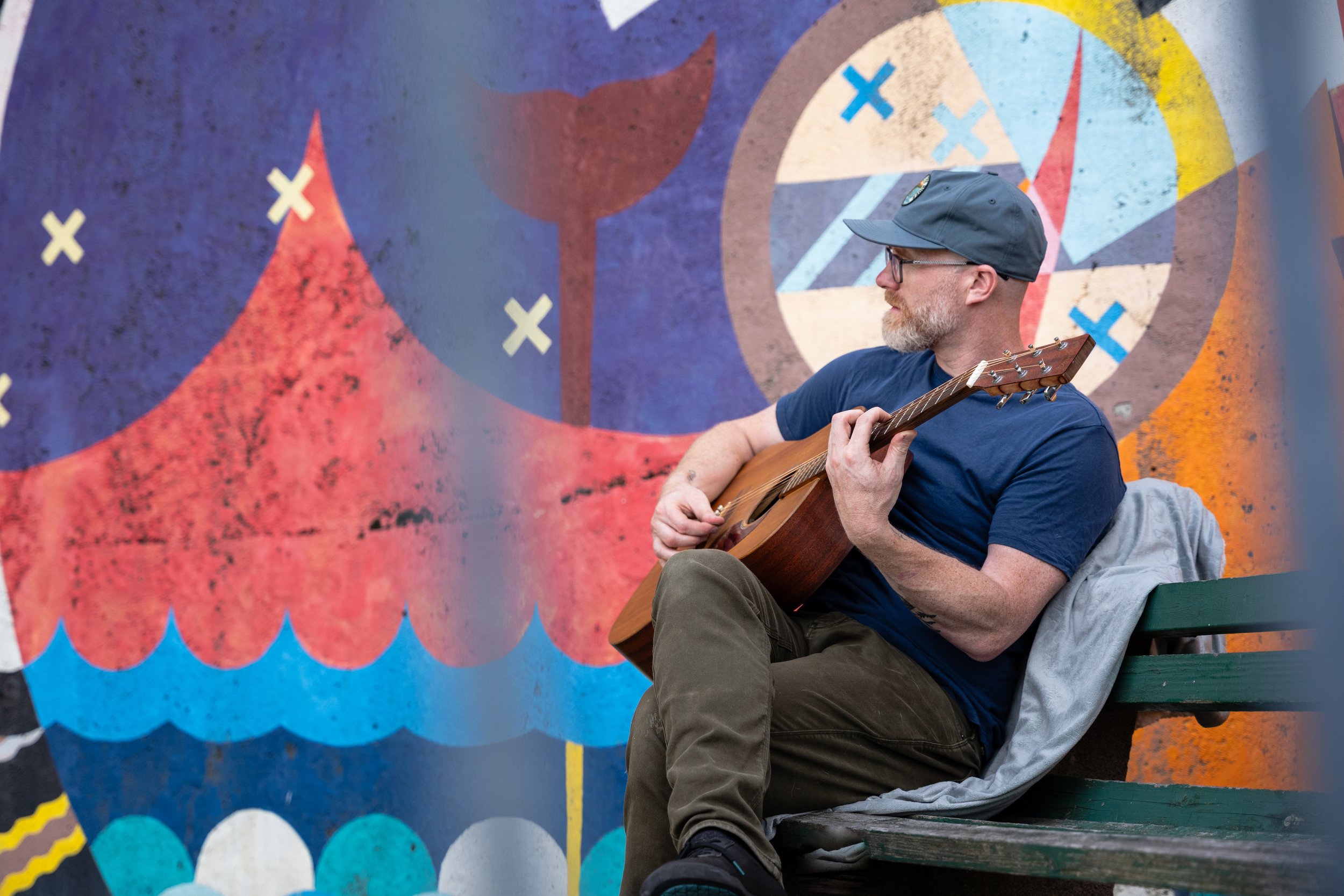 A person wearing glasses, a gray cap, and a blue shirt is playing an acoustic guitar while sitting on a bench outdoors. He is positioned in front of a colorful abstract mural with geometric shapes and bright colors.