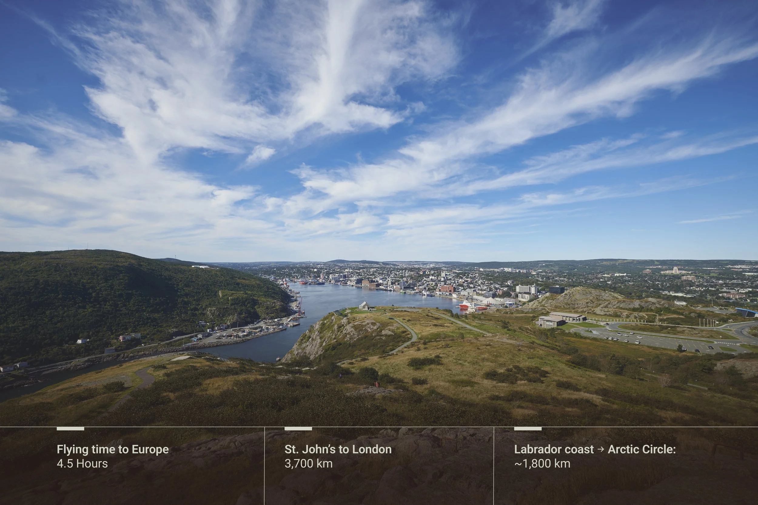 A panoramic view of St. John's harbour with ships, surrounded by green hills and a cityscape under a blue sky with clouds.
