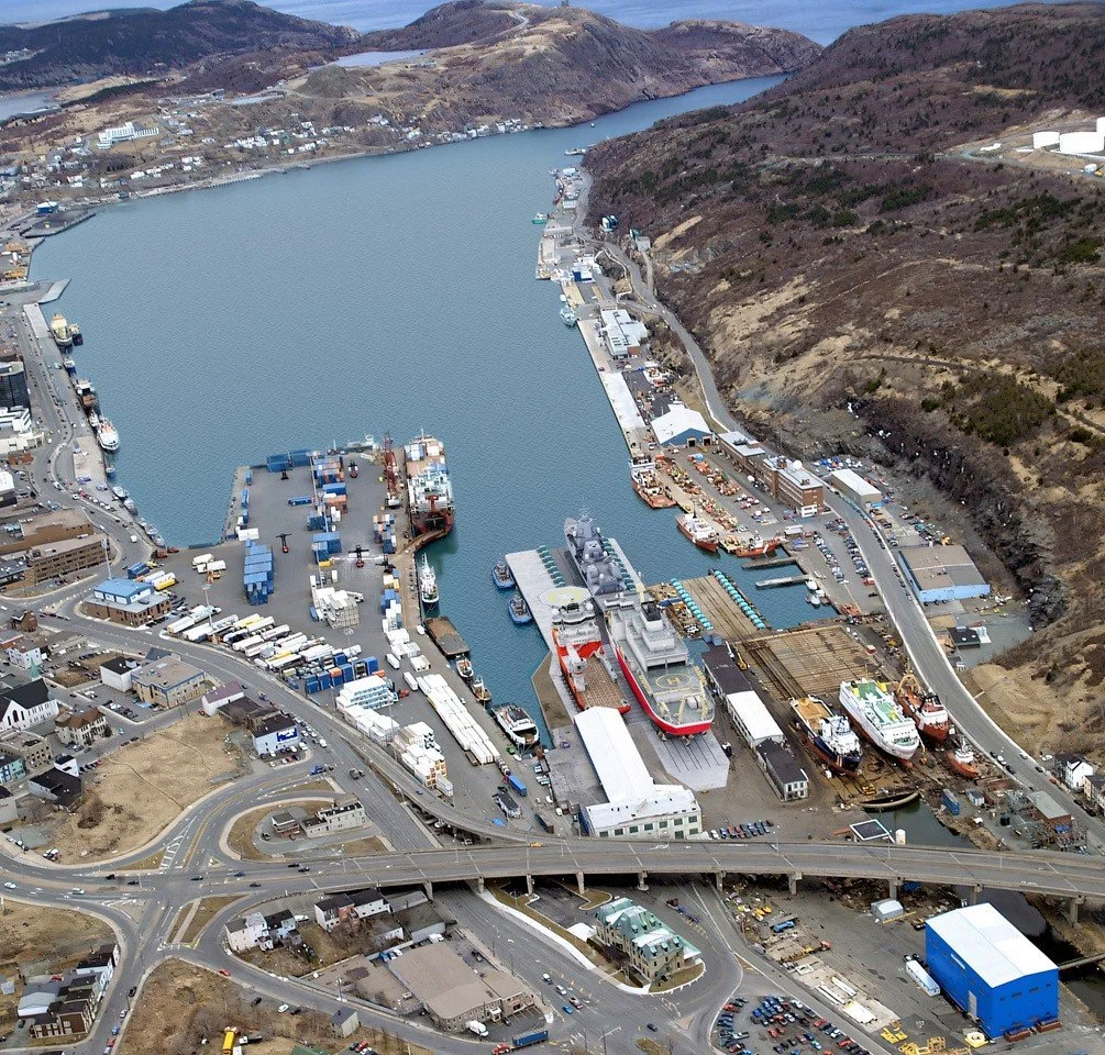 Aerial view of a harbour port with ships and industrial buildings, with roads and a bridge in the foreground, and hills in the background.