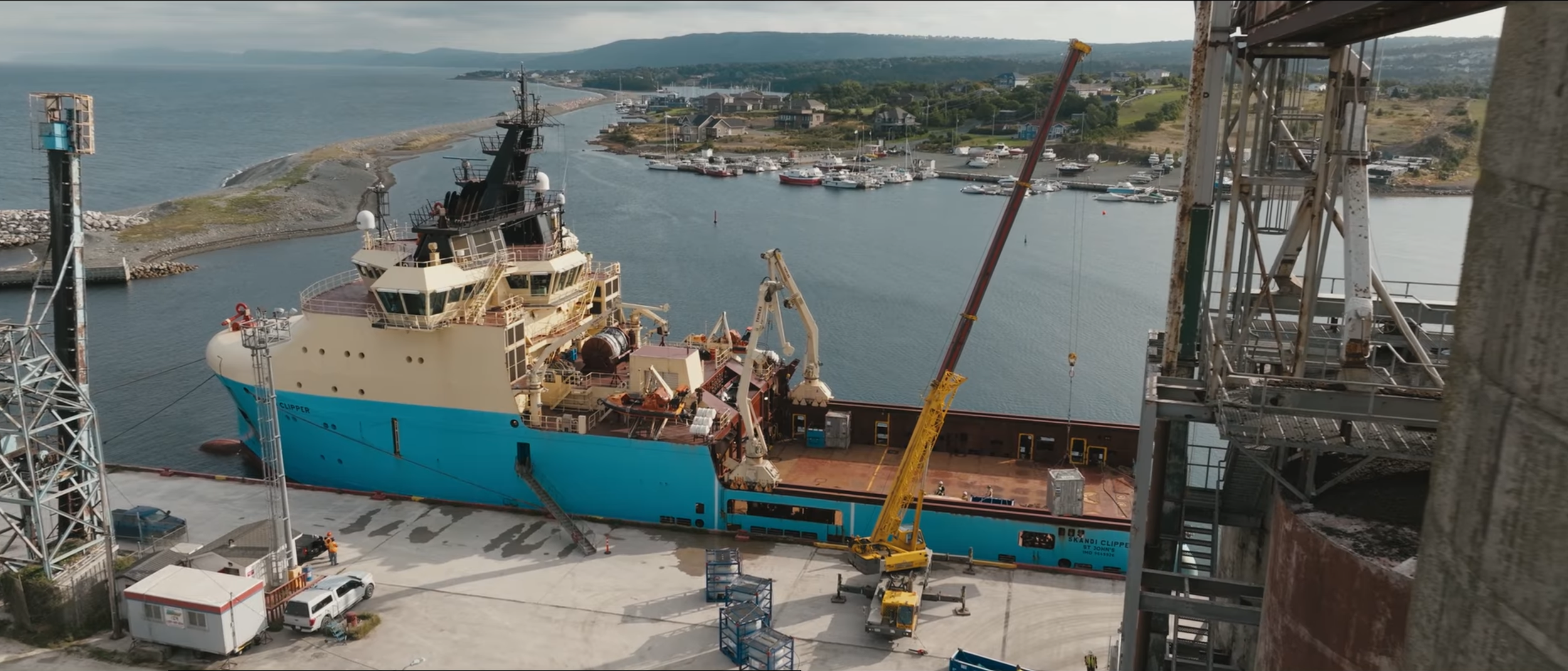 Large ship docked at a shipyard with construction cranes and scaffolding, with smaller boats and water in the background.